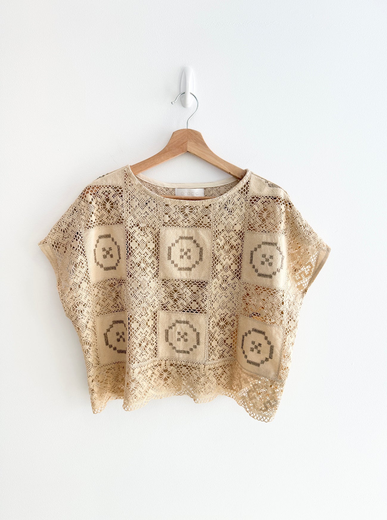 Beige crochet top with geometric patterns on a wooden hanger against a white background