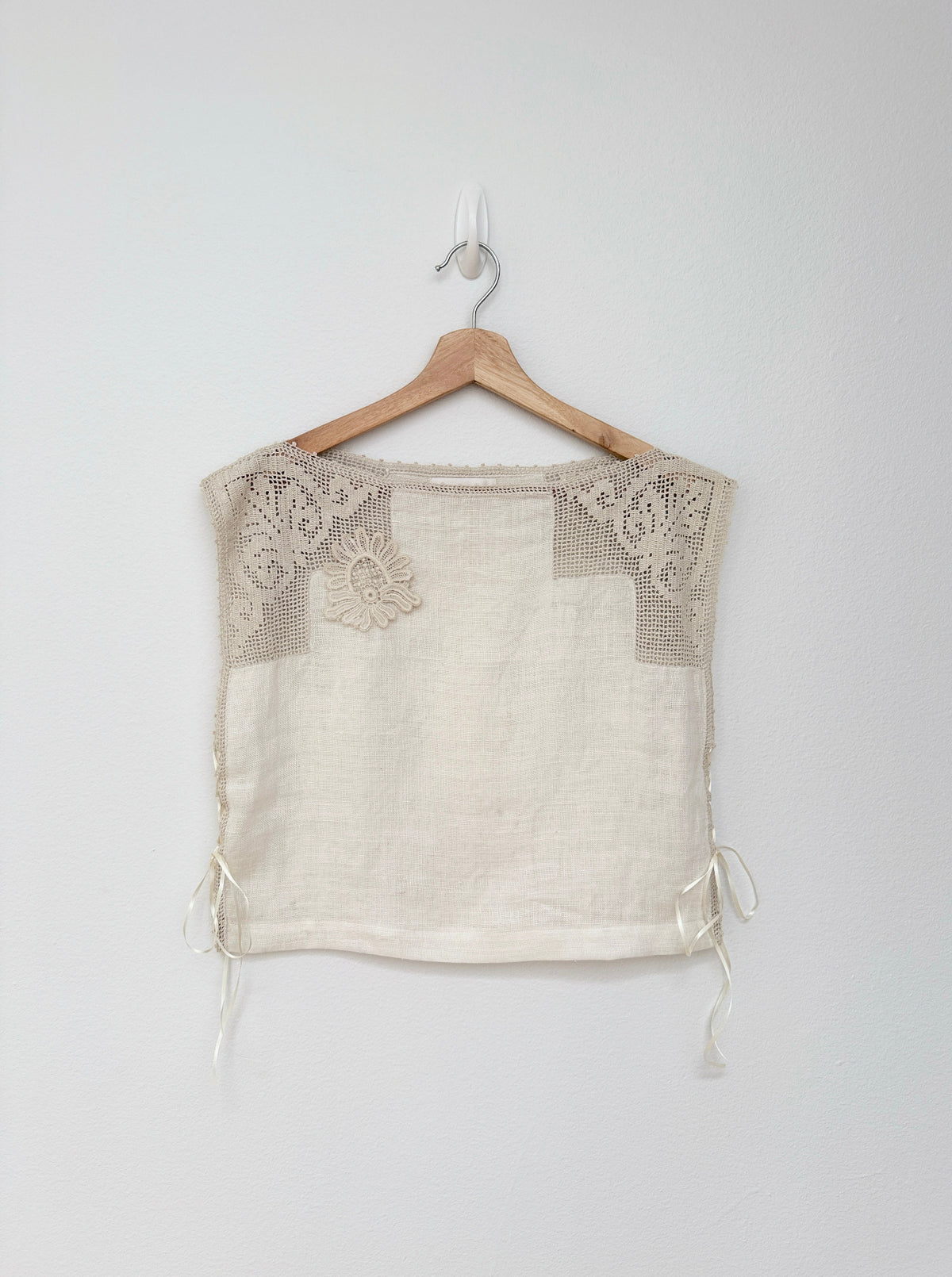An off-the-shoulder cream linen top with beige crochet lace detailing on the chest and side ties, hanging on a wooden hanger against a light gray wall.