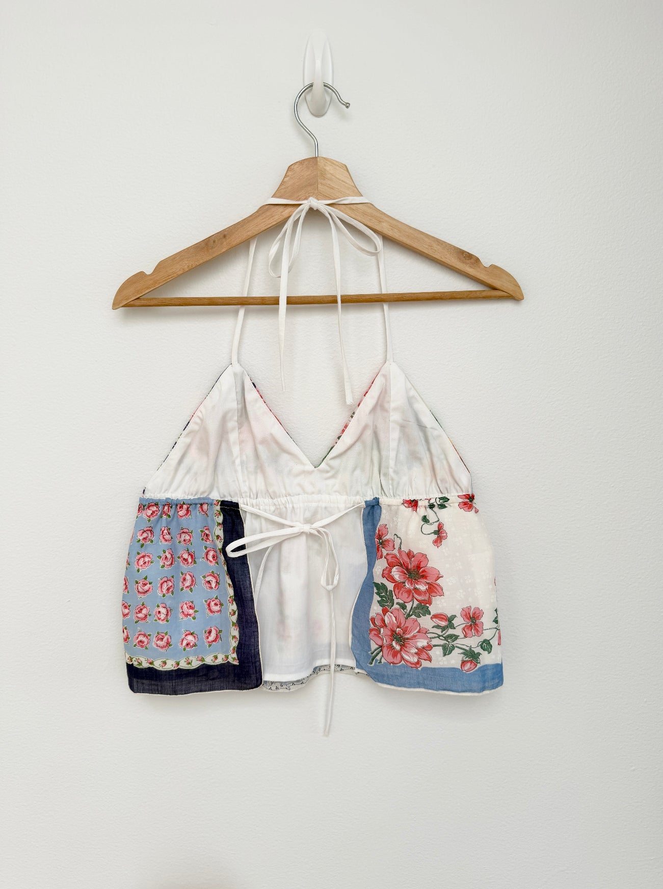 A white halter crop top with mixed floral and patterned fabric panels, hanging on a wooden hanger against a plain white wall.