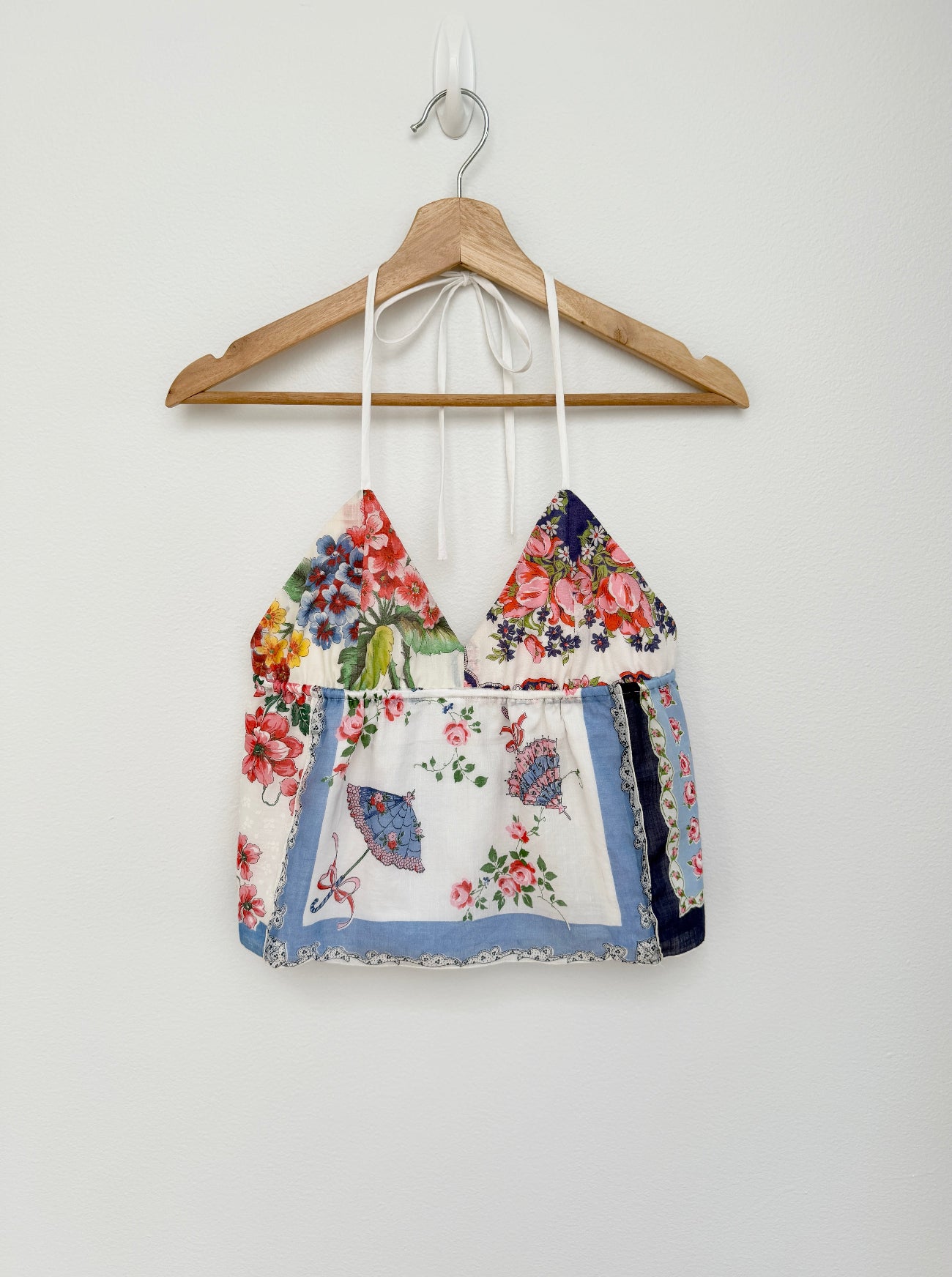 A halter-style crop top with a multicolor floral and butterfly print, hanging on a wooden hanger against a plain white wall.