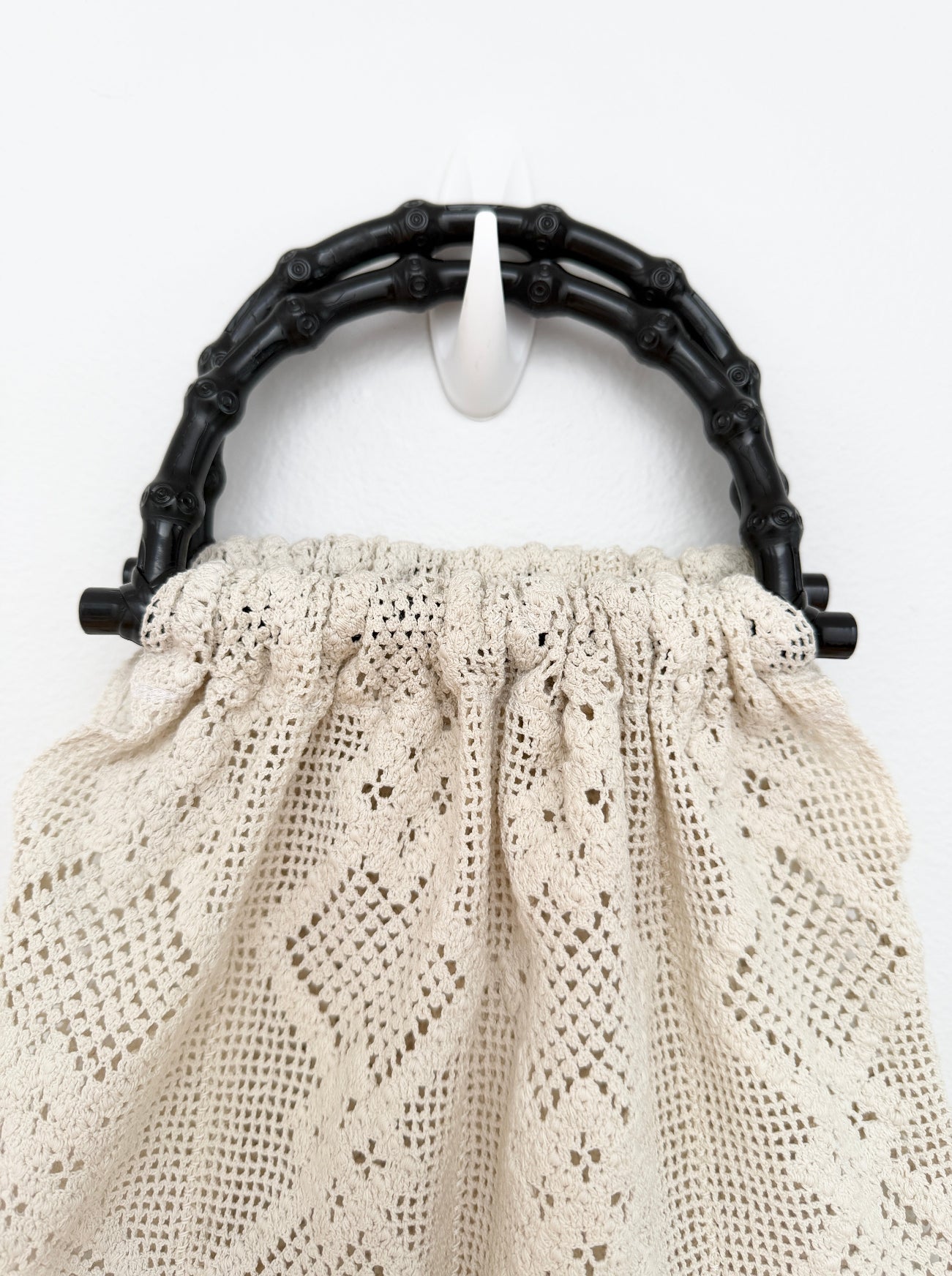 A close-up of a cream-colored crochet or lace handbag with a black bamboo-style top handle, shown against a plain white background.