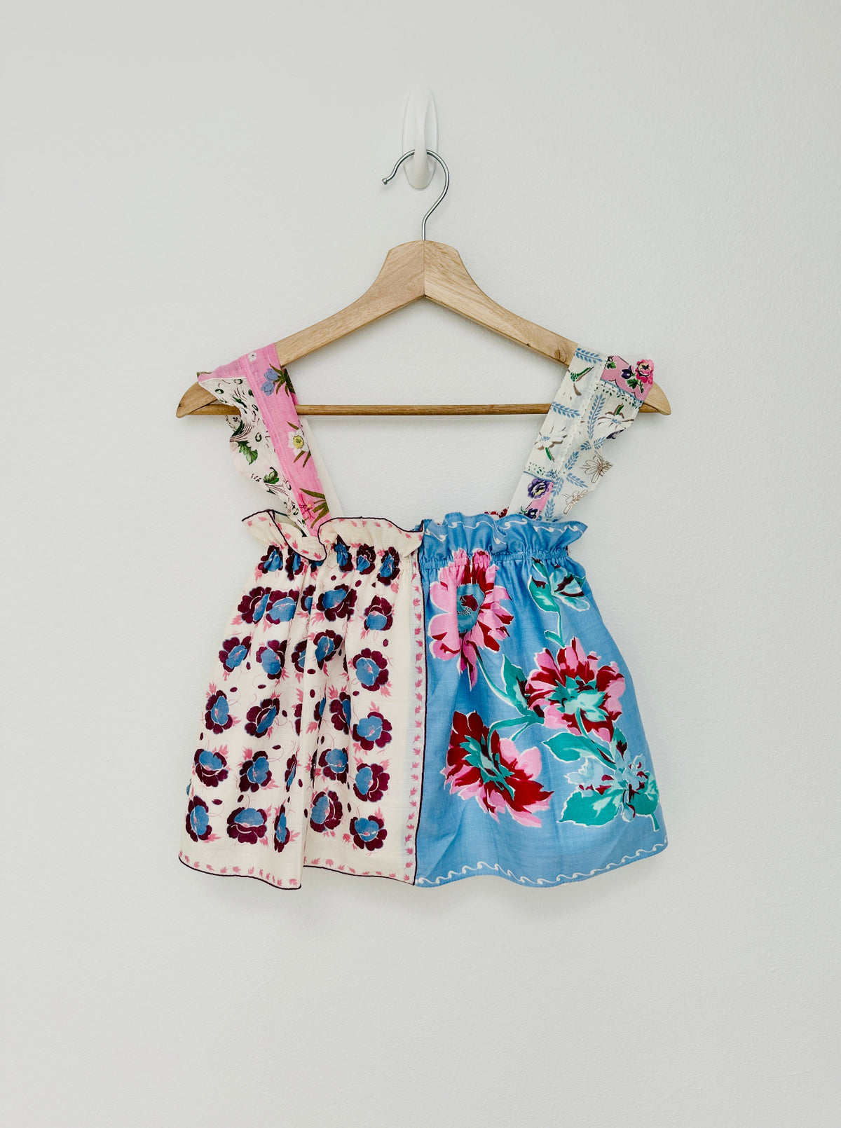 A women's sleeveless top with a split design hangs on a wooden hanger against a plain light gray wall. The left side is light pink with dark floral motifs, and the right side is light blue with large pink, red, and teal floral prints.