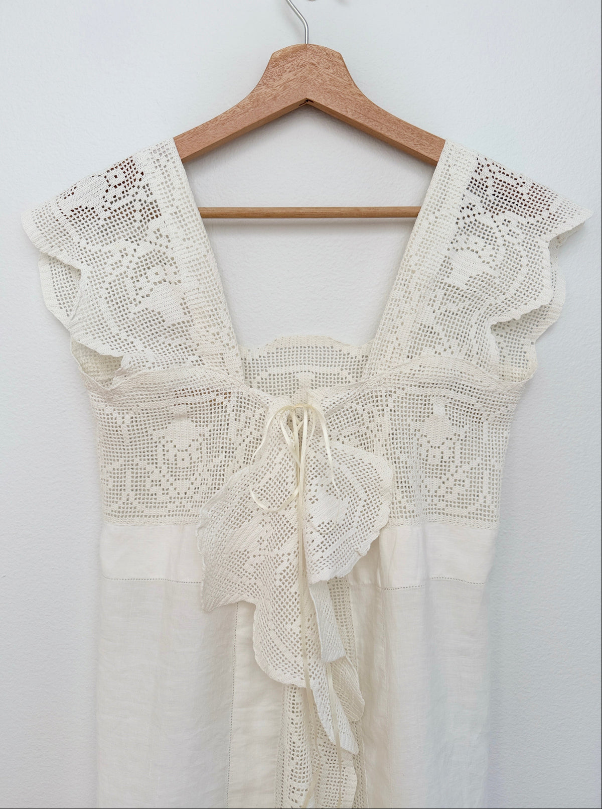An off-white crochet dress hanging on a wooden hanger against a plain white wall. The dress features a square neckline with wide ruffled lace shoulder panels, a matching lace bodice with a back tie, and a tiered skirt.