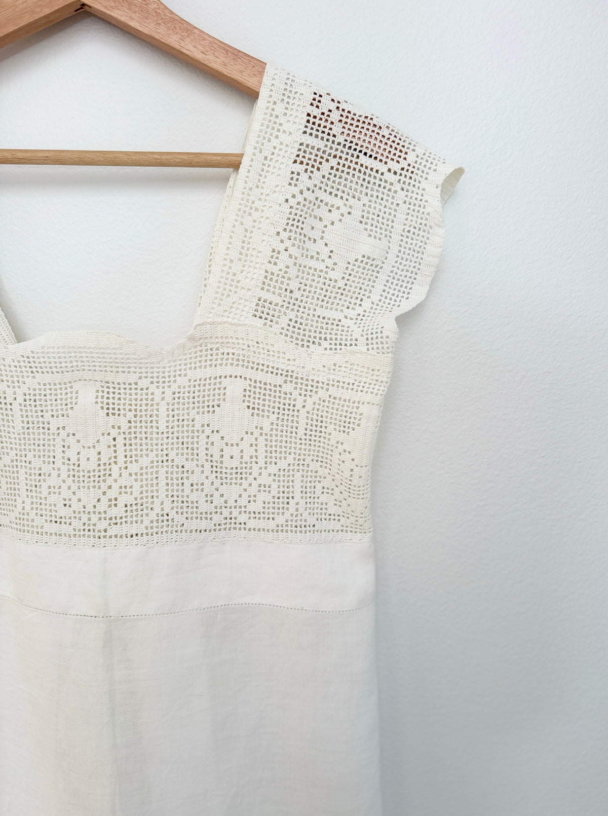 An off-white sleeveless dress with intricate floral lace embroidery across the bodice and shoulder, hanging on a wooden hanger against a plain white wall.