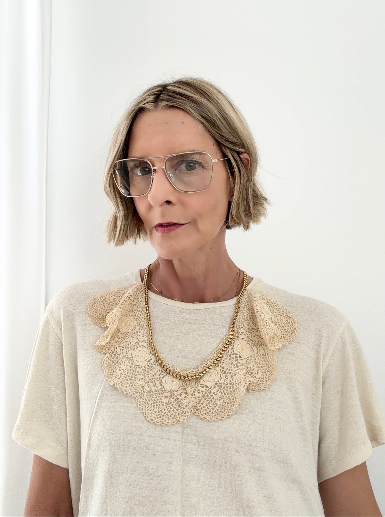 Woman wearing a beige blouse with a lace collar and gold necklace against a white background