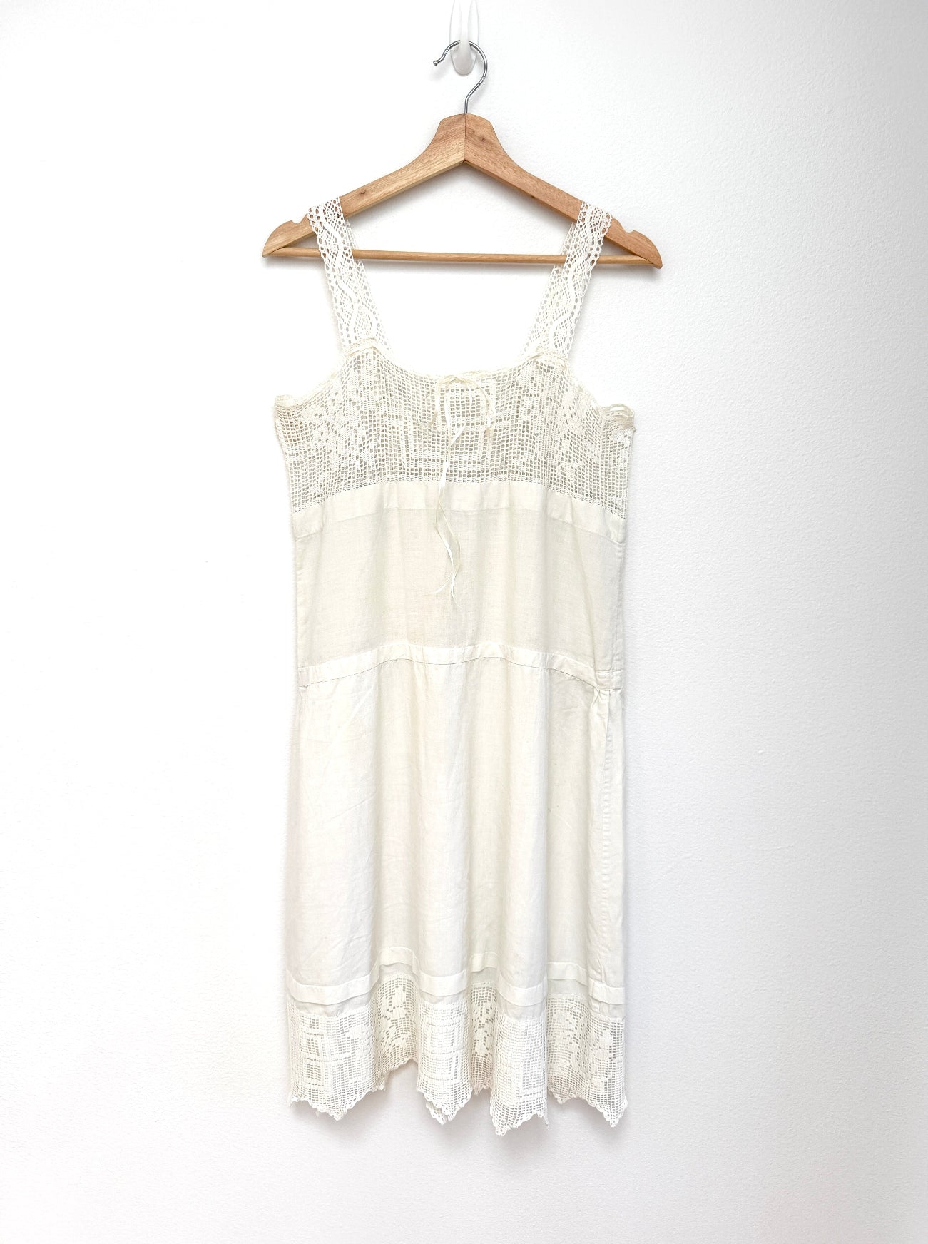 An off-white sleeveless lace and embroidered nightgown or dress hanging on a wooden hanger against a plain white wall.