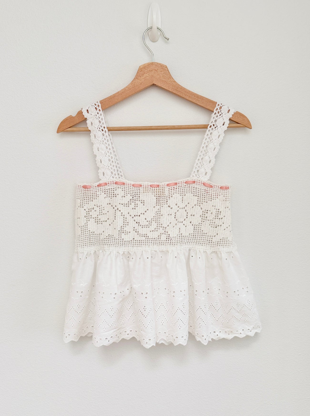 White eyelet lace sleeveless top with floral lace bodice and ruffled hem, hanging on a wooden hanger against a light gray wall.