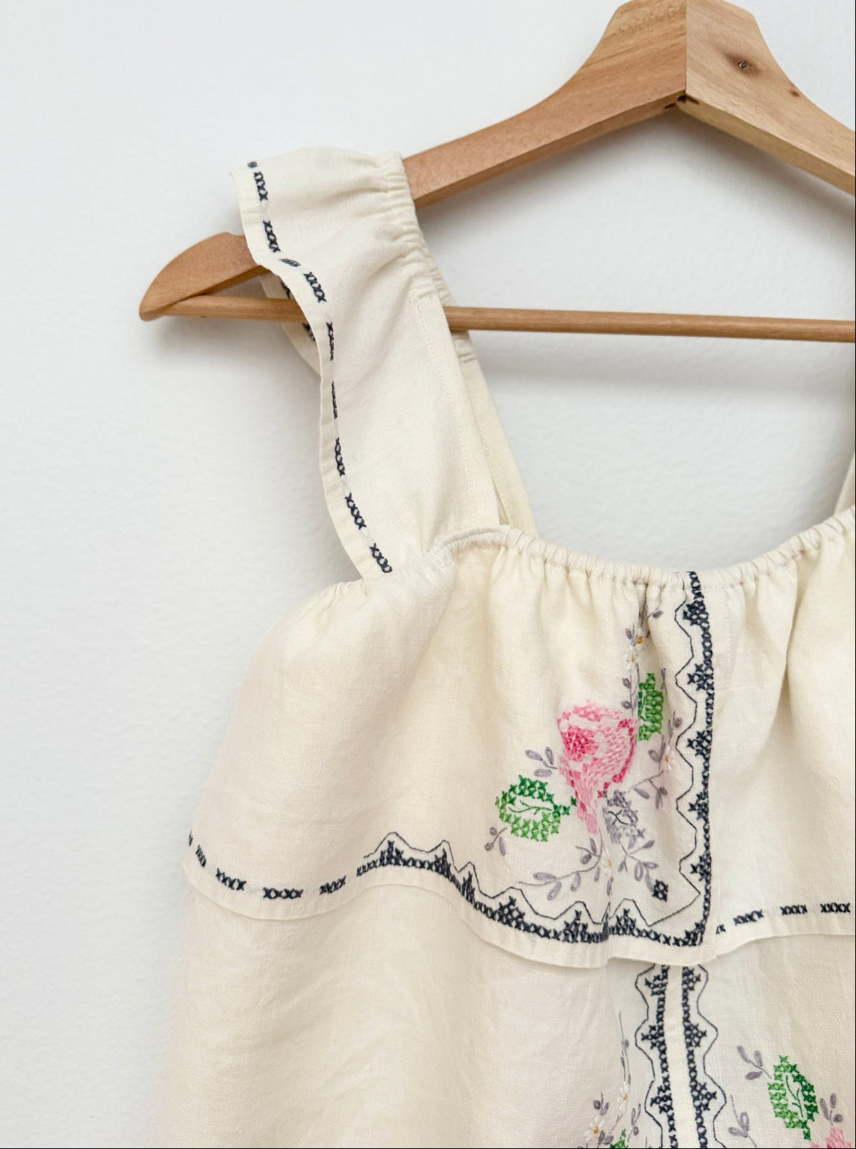 An off-white cotton top with pink and green floral embroidery and black patterned trim, hanging on a wooden hanger against a white wall.
