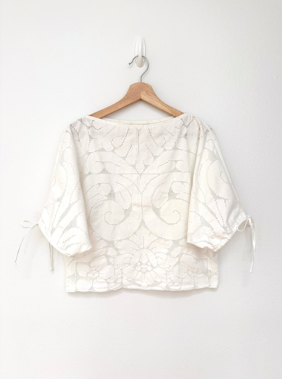A white lace blouse with short puff sleeves and side tie details, hanging on a wooden hanger against a plain white wall.