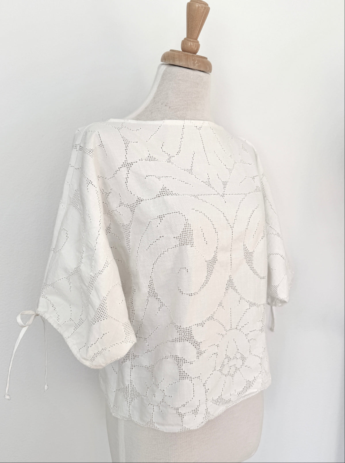 A white floral lace top displayed on a dress form mannequin against a plain white wall. The top features short sleeves with side ties and a sheer embroidered pattern.