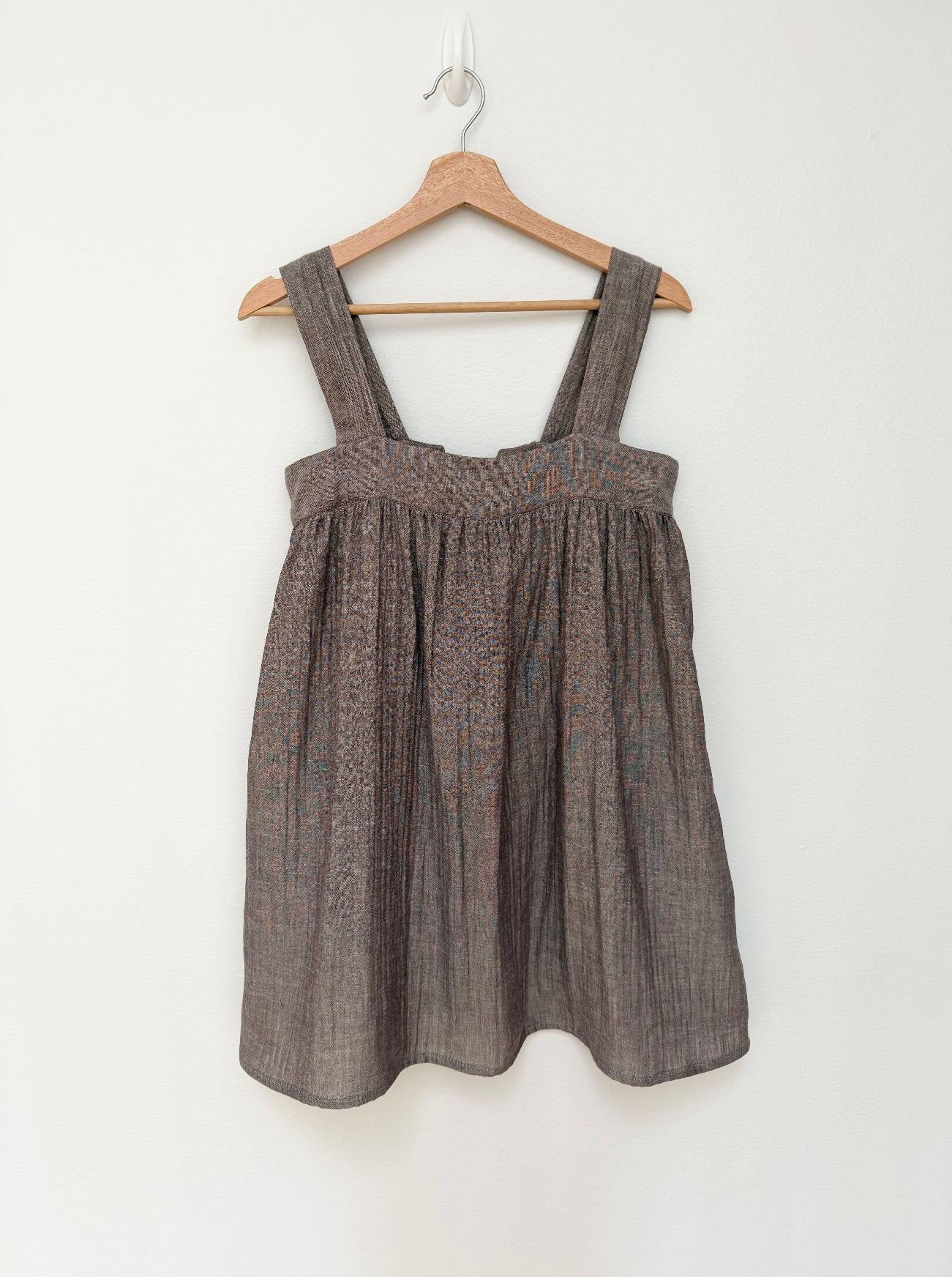 A sleeveless brown textured linen dress with wide shoulder straps, hanging on a wooden hanger against a plain white wall.