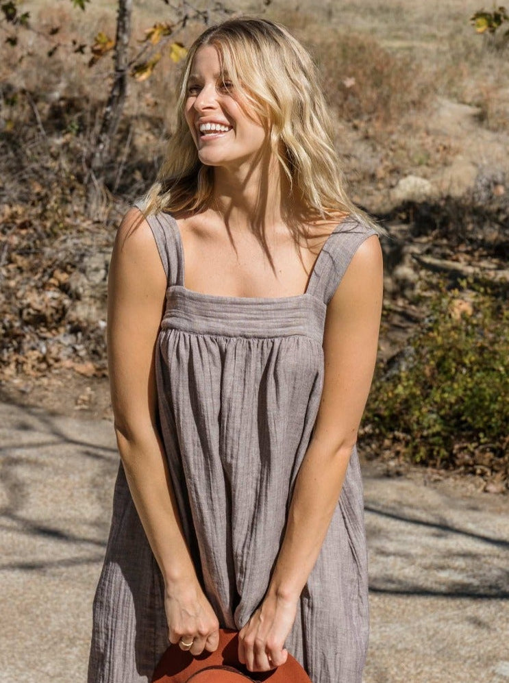 A woman wearing a sleeveless, midi-length gray linen dress stands outdoors on a stone path holding a rust-colored hat.