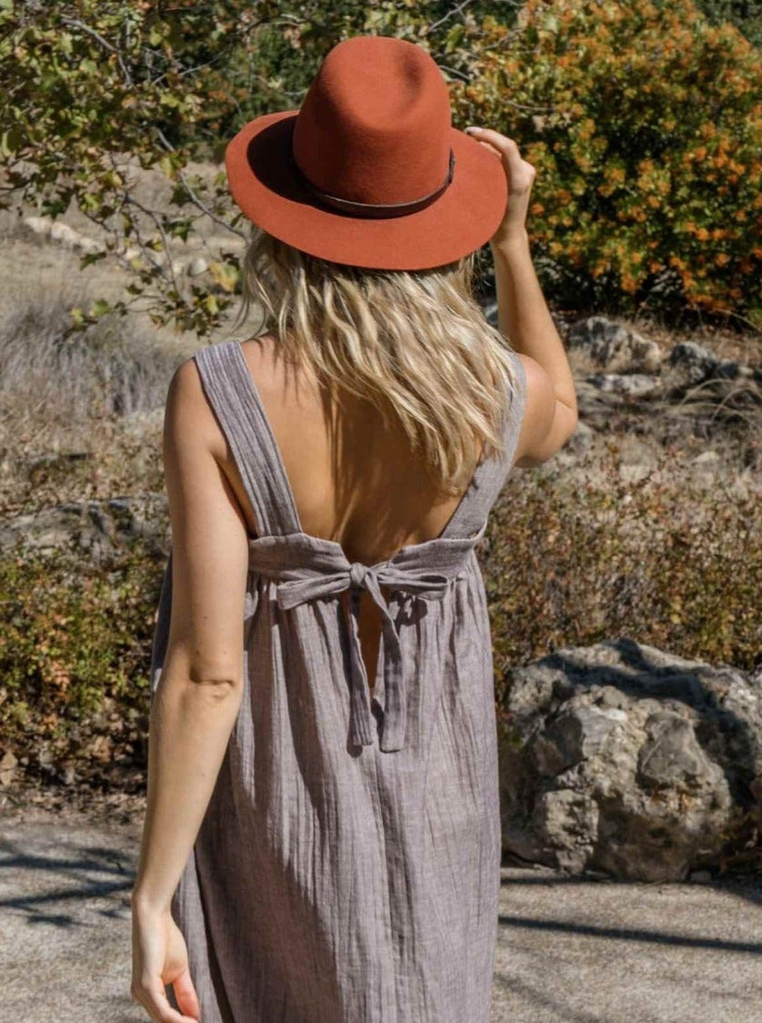 A woman viewed from behind wearing a sleeveless, backless midi dress in a muted taupe-gray color and a rust-orange fedora hat, standing outdoors on a stone path with trees and shrubs in the background.