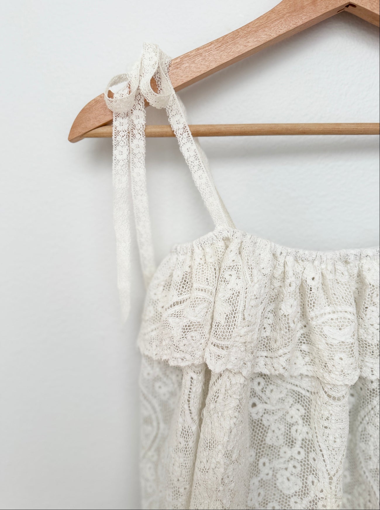 An off-white lace halter top with ruffled trim hanging from a wooden hanger against a plain white wall.