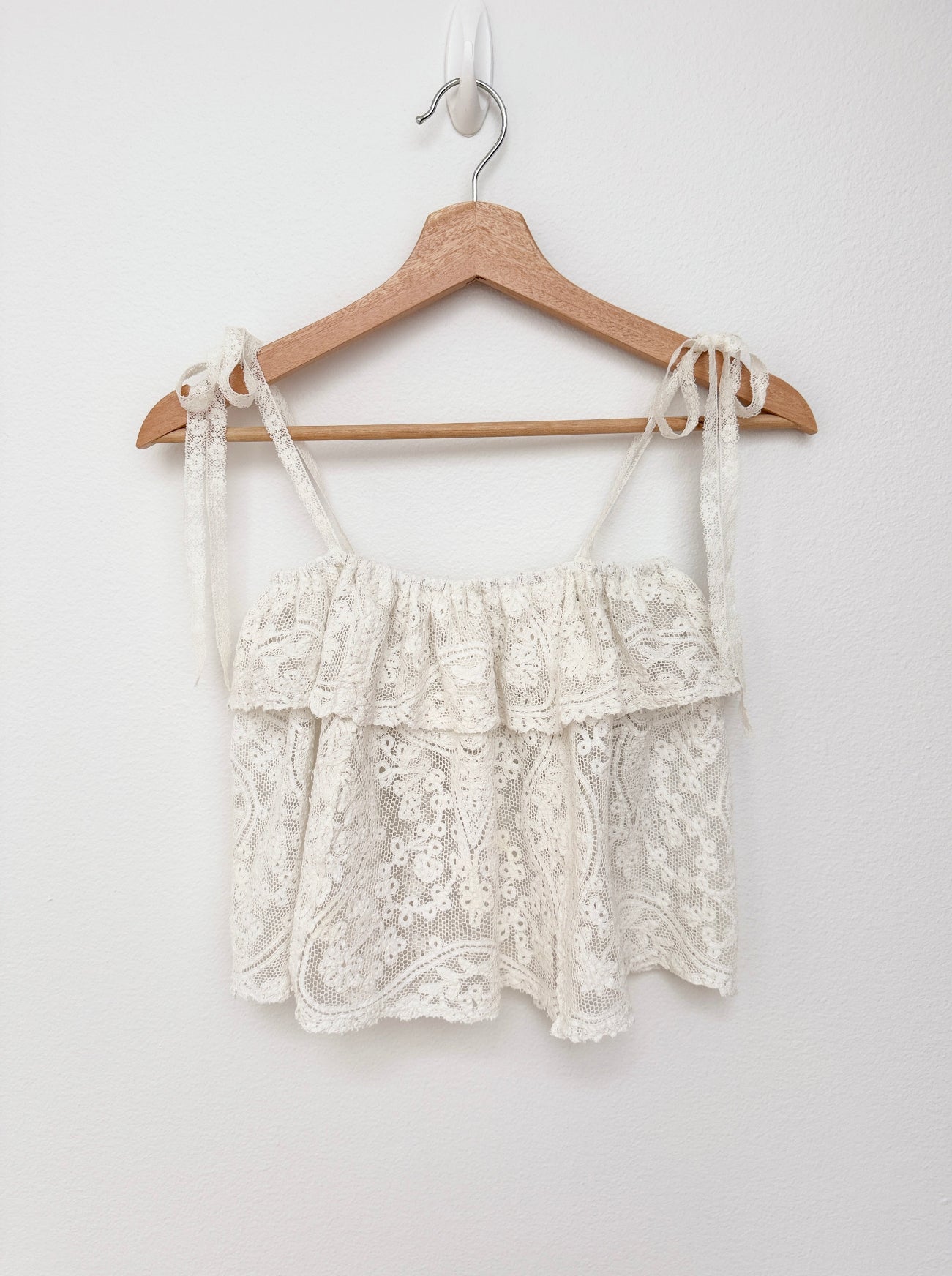 An off-white lace camisole top with tie straps, hanging on a wooden hanger against a plain white wall.
