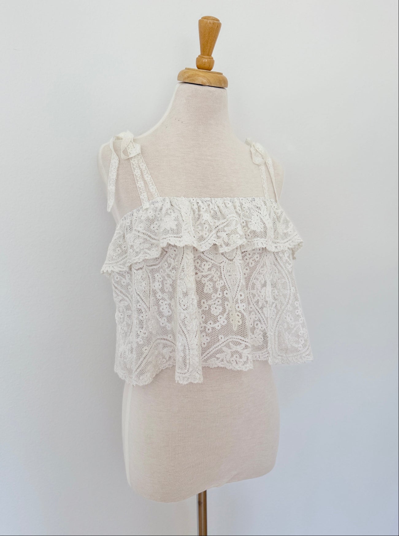 White lace ruffle cropped tank top displayed on a cream fabric dress form mannequin against a plain white wall.