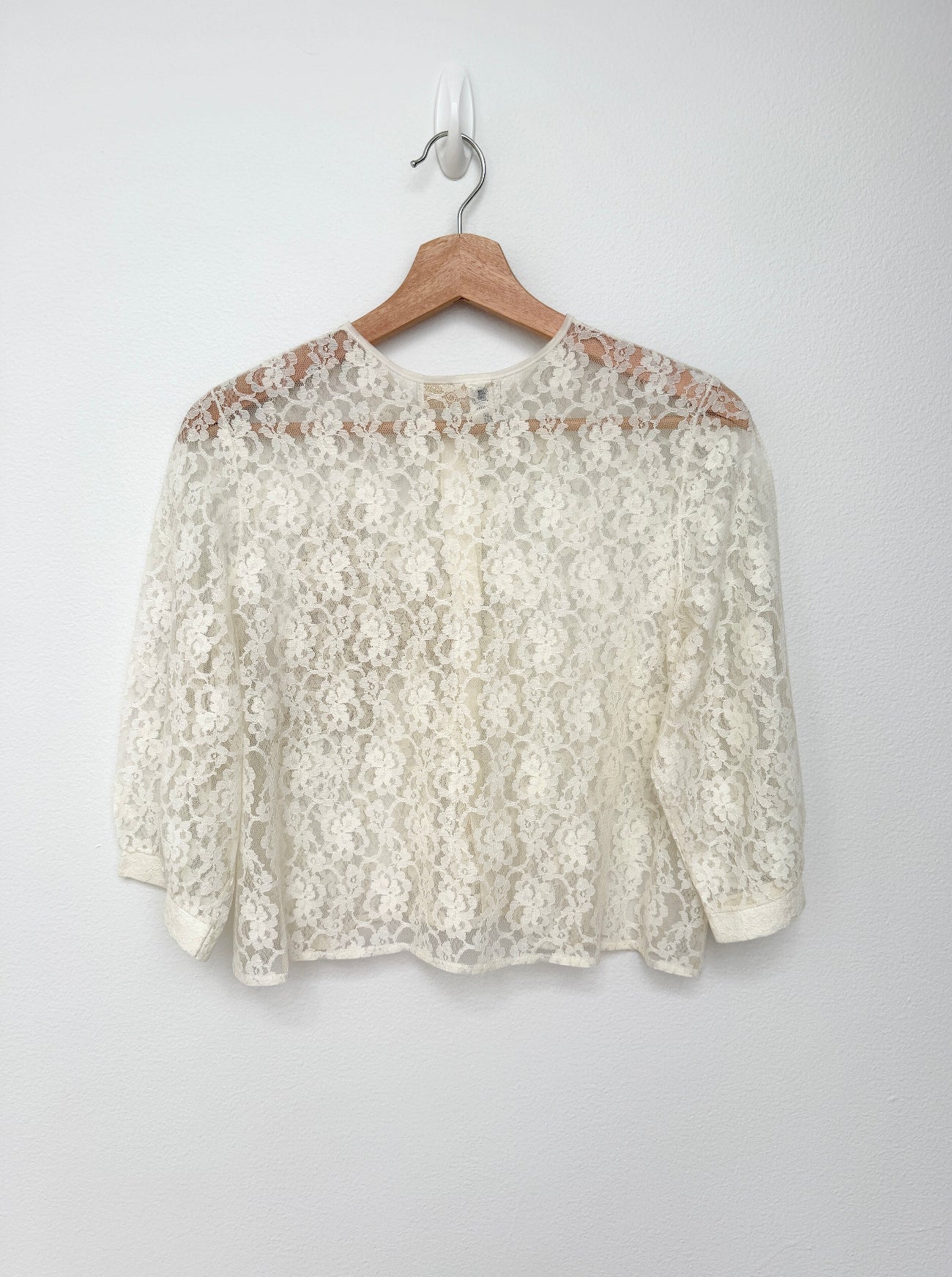 An off-white floral lace long-sleeve top hanging on a wooden hanger against a plain light gray wall.