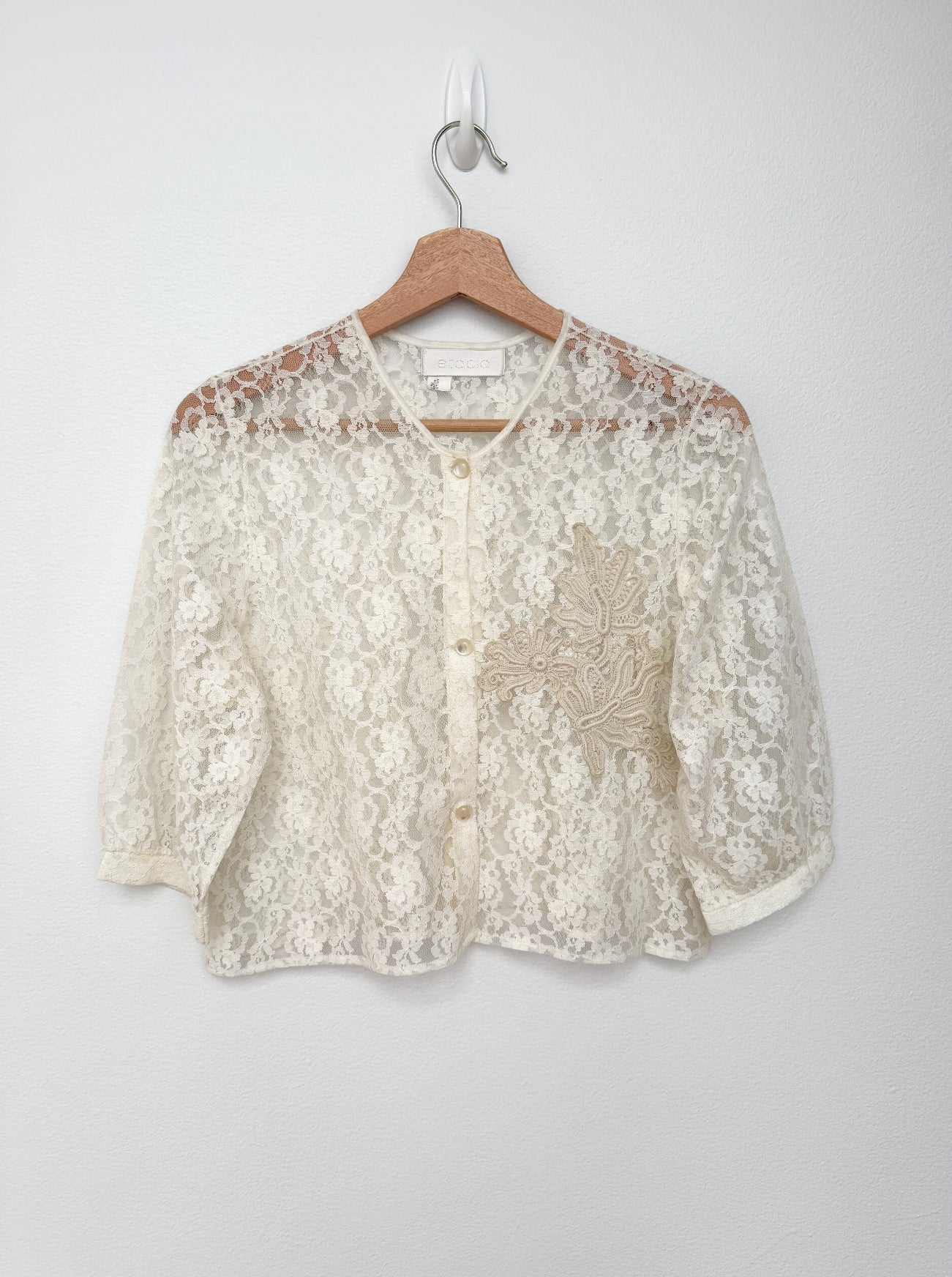 An off-white lace cardigan with floral embroidery on the front, hanging on a wooden hanger against a plain light gray wall.