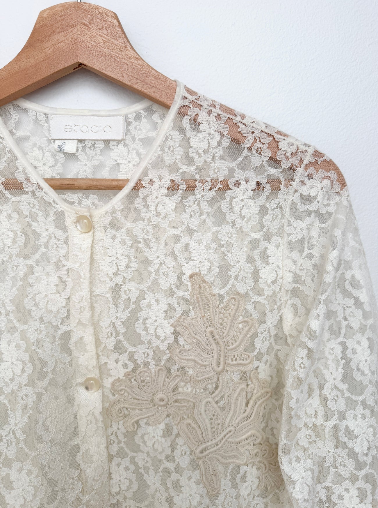 An ivory lace blouse with floral embroidery and a buttoned neckline, hanging on a wooden hanger against a white wall. The brand name 'etcio' is visible on the inner neck label.