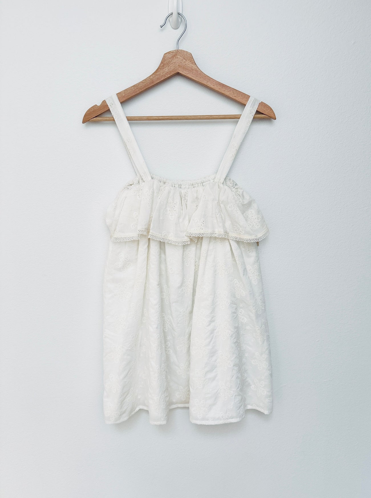An off-white textured cotton sleeveless mini dress with ruffled layers and thin shoulder straps, hanging on a wooden hanger against a plain light gray wall.