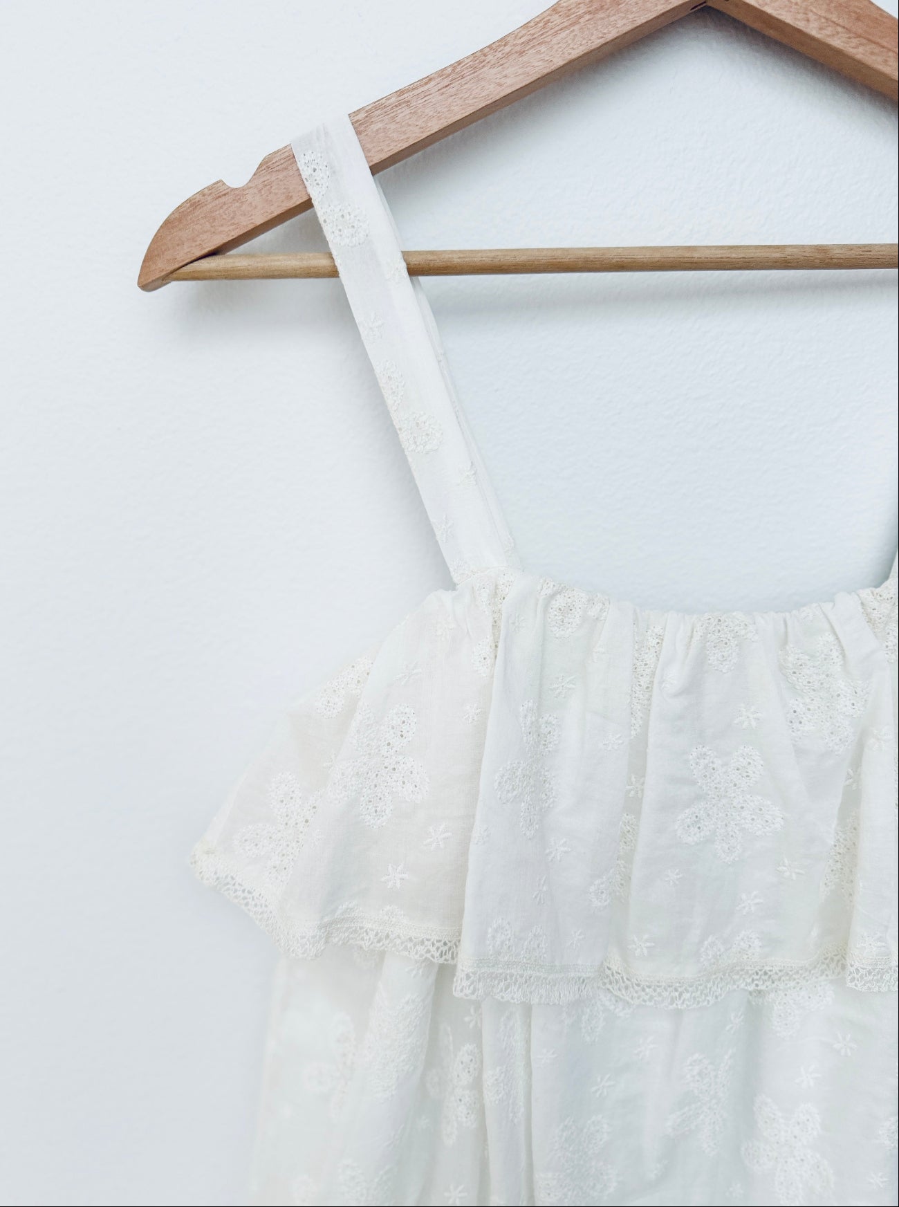 White embroidered tiered dress with thin shoulder straps hanging on a wooden hanger against a white wall.