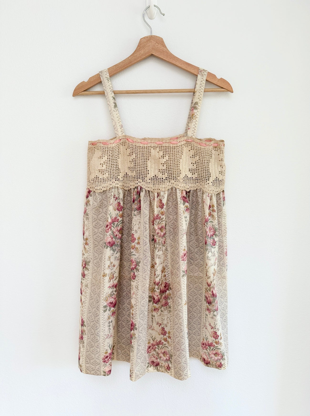 A sleeveless babydoll dress with a beige and brown floral print and a textured crochet lace bodice, hanging on a wooden hanger against a plain white wall.