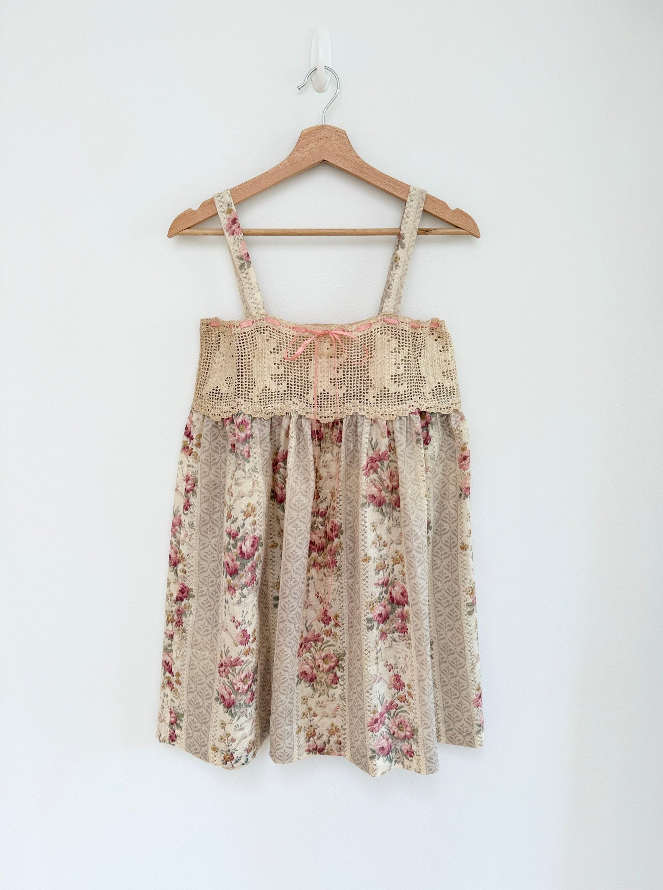A sleeveless babydoll dress with a beige lace bodice and a beige floral skirt, hanging on a wooden hanger against a plain white wall.