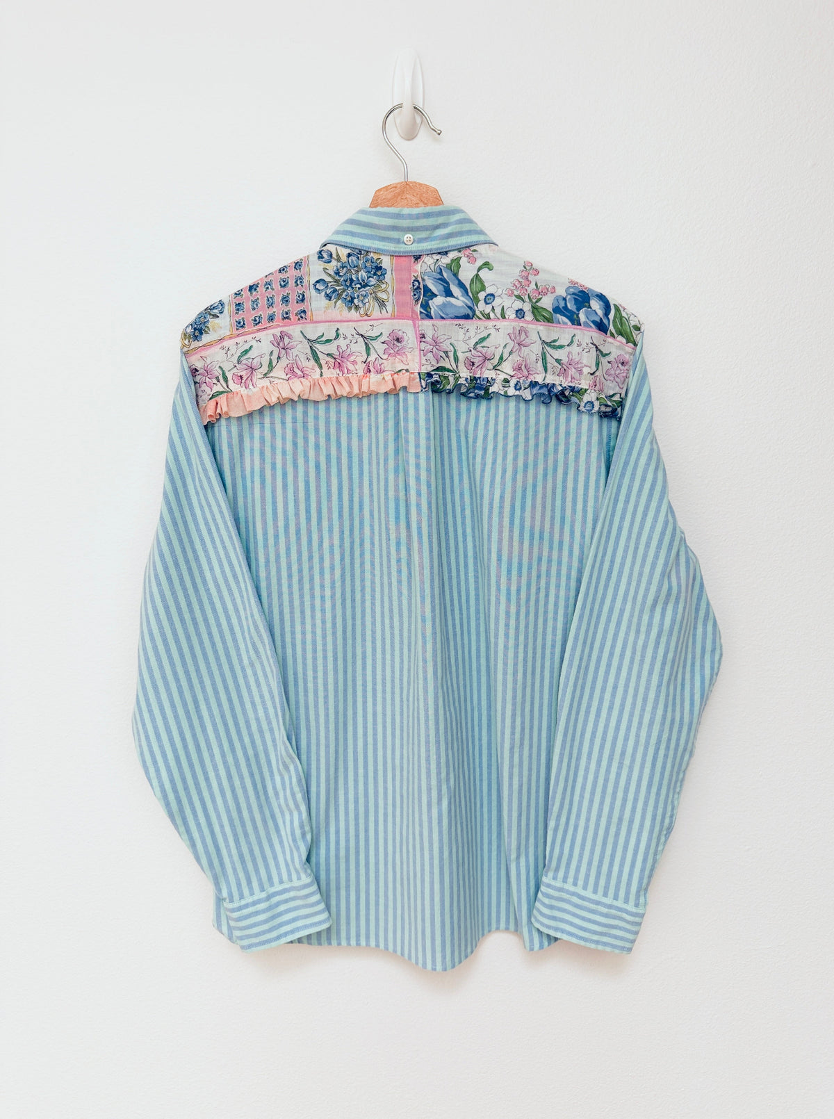 A light blue and white vertically striped long-sleeve shirt with a colorful floral and patterned yoke across the shoulders, hanging on a wooden hanger against a plain white wall.