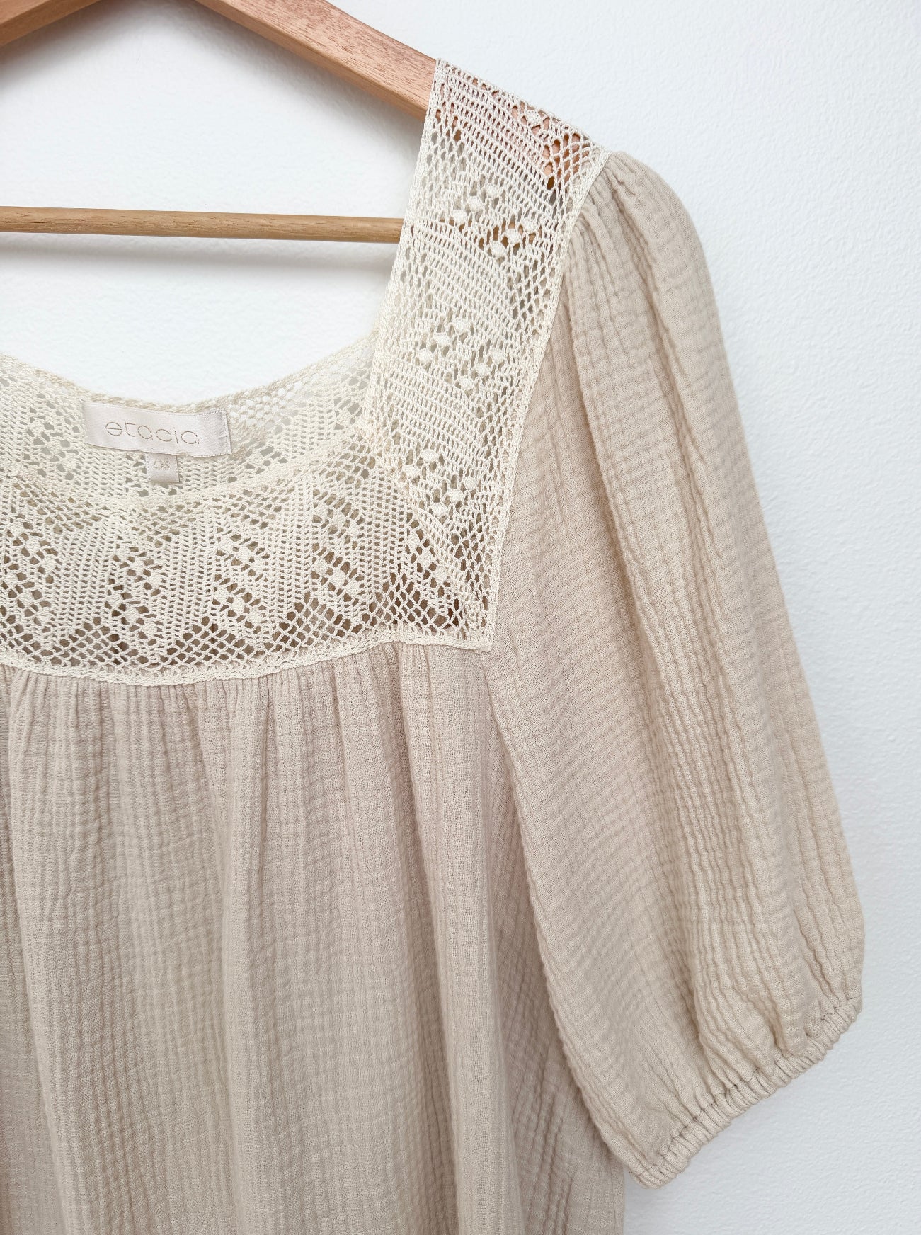 A close-up of a beige gauze mini dress hanging on a wooden hanger against a white wall. The blouse features a square neckline with a cream-colored crochet lace panel and gathered puff sleeves.