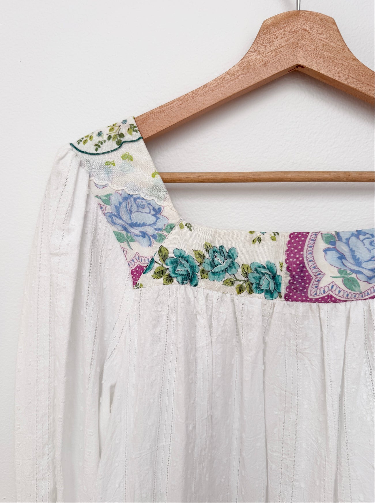 White textured cotton dress with a floral printed yoke featuring blue roses and green leaves, hanging on a wooden hanger against a white wall.