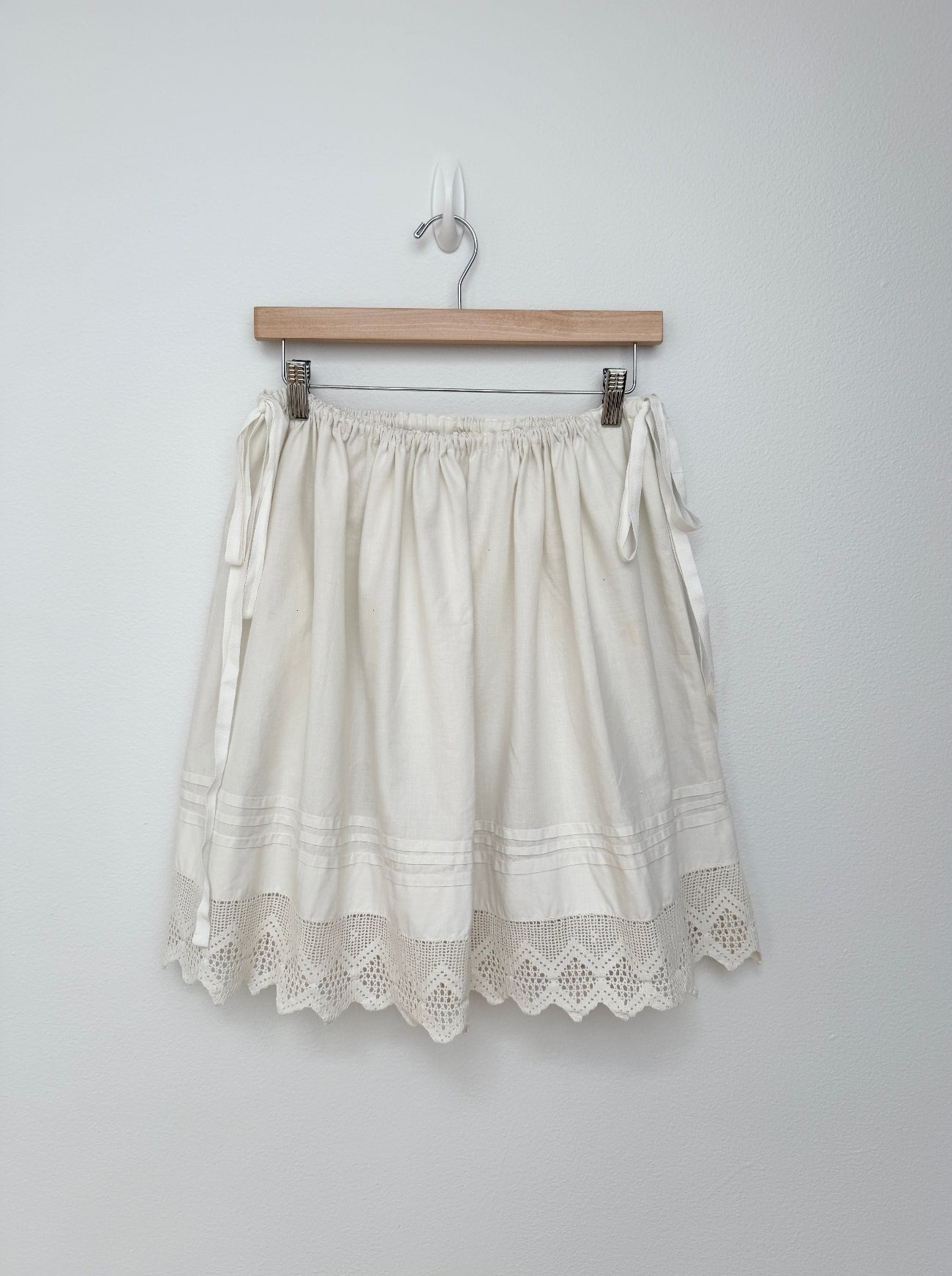 An off-white cotton skirt with embroidered details and a scalloped eyelet lace hem, hanging from a wooden hanger against a plain light gray wall.