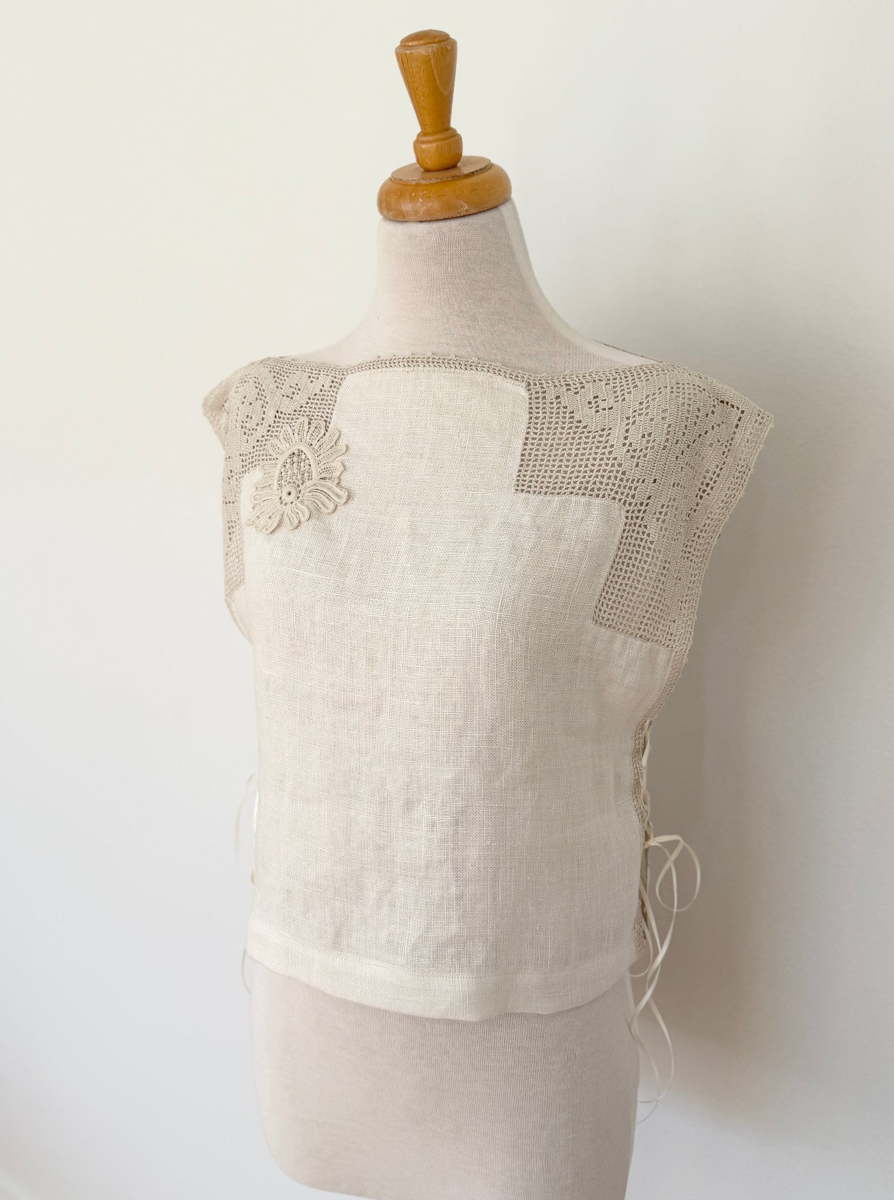 A cream-colored linen vest with floral lace applique and crochet shoulder panels, displayed on a beige fabric dress form mannequin against a plain white wall.