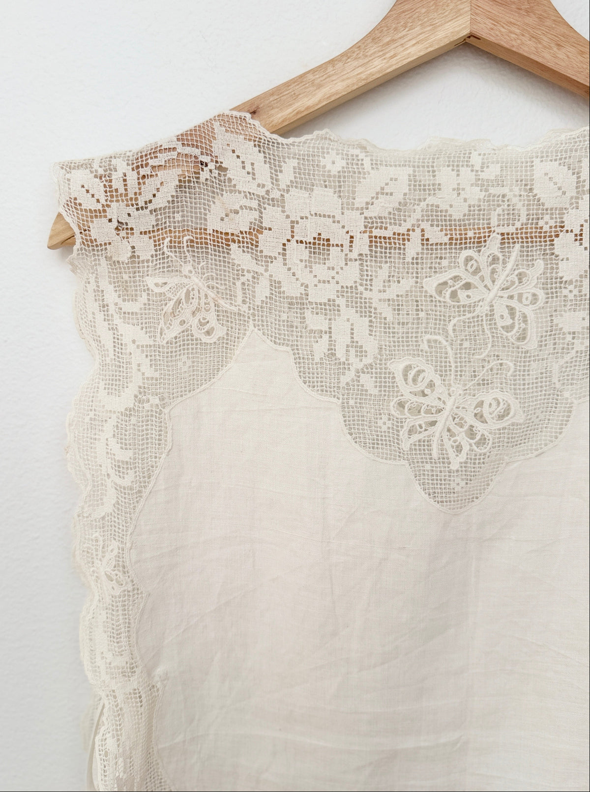 An off-white lace and linen textile with a floral embroidered lace border, hanging on a wooden hanger against a white wall.