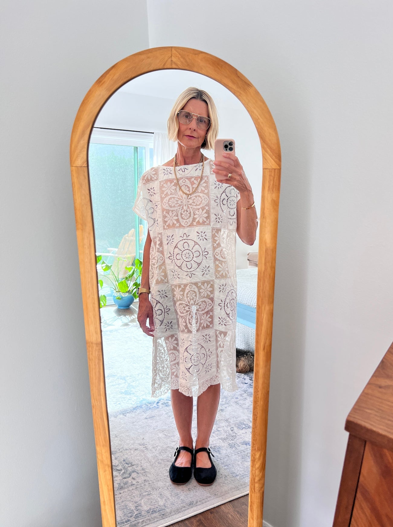 Woman taking a mirror selfie wearing a white lace dress in a room with a wooden floor.