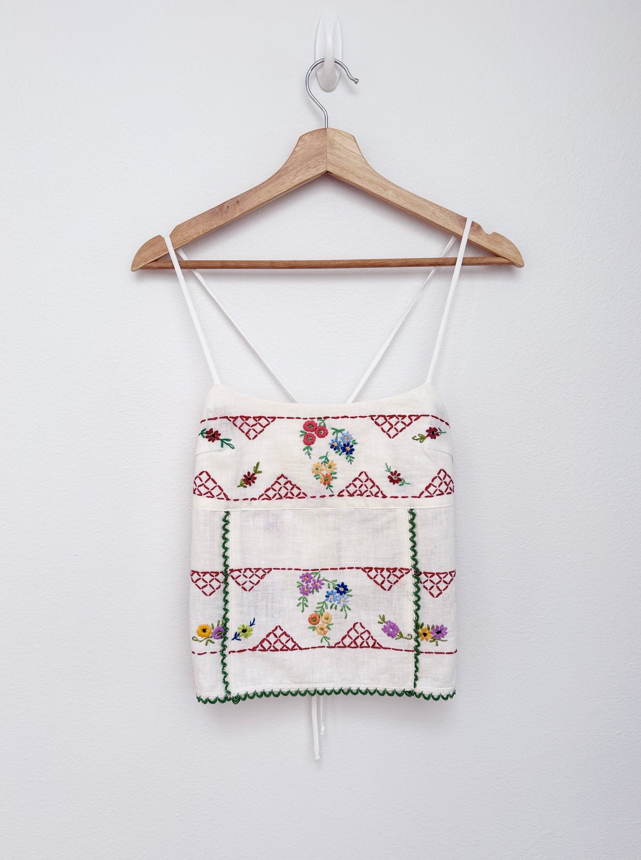 An embroidered white camisole top with red geometric patterns and multicolor floral embroidery, hanging on a wooden hanger against a plain white wall.