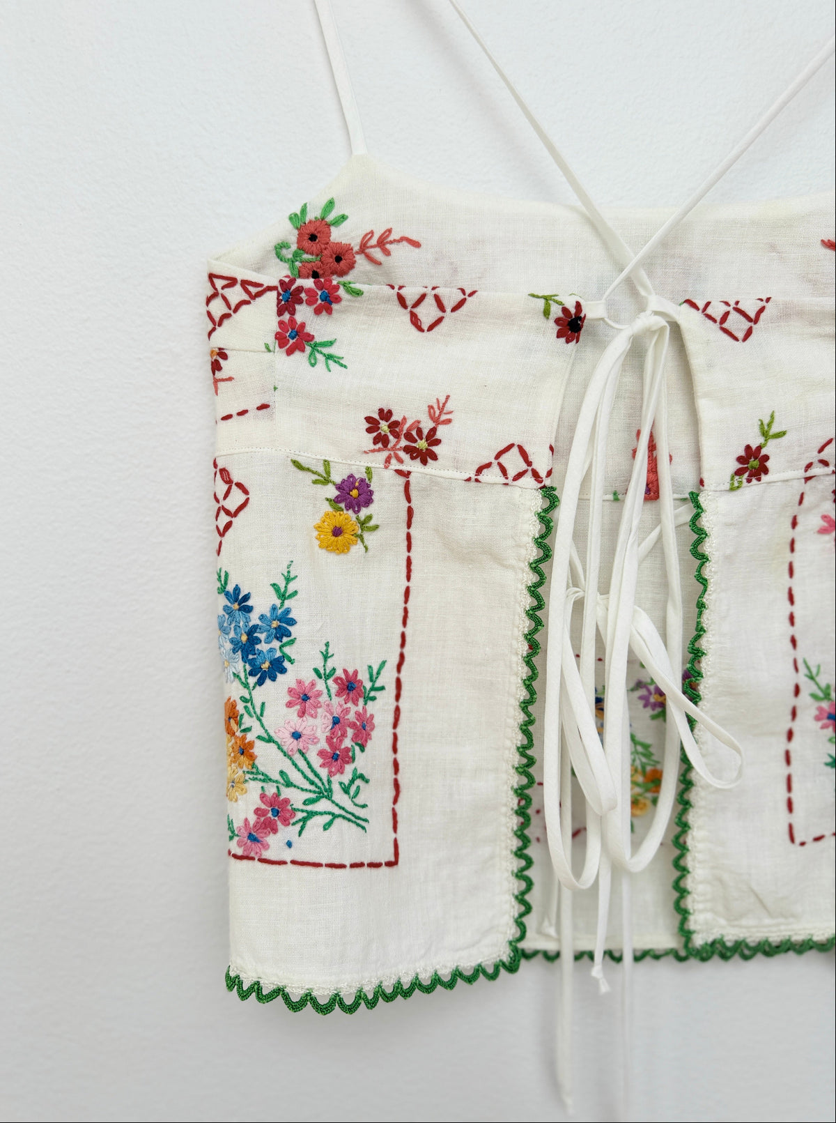 A close-up of a white embroidered fabric garment, likely a blouse or apron panel, featuring colorful floral embroidery, red stitched borders, green scalloped edges, and white drawstrings against a plain white background.