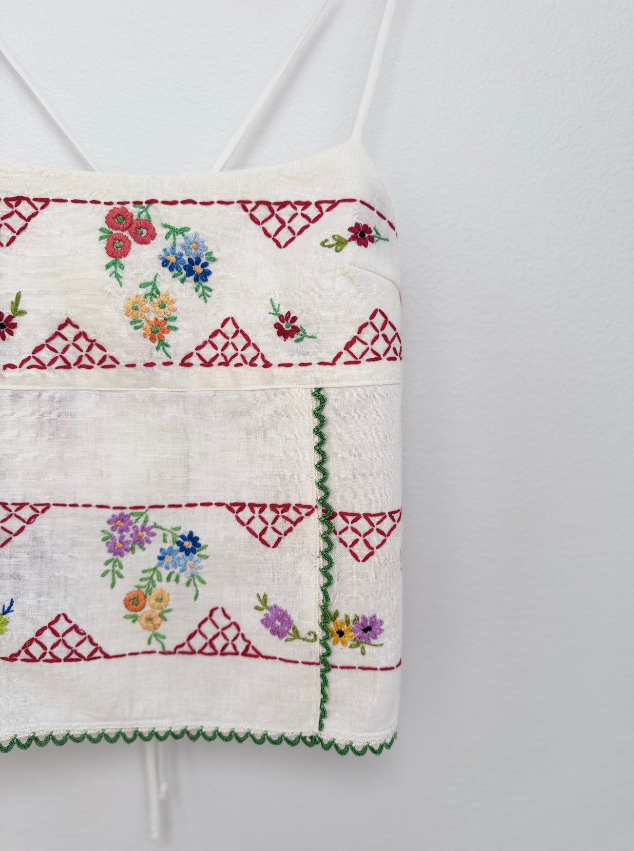 A close-up of a white embroidered fabric garment, likely a strap top or skirt panel, featuring red geometric borders, green zigzag trim, and multicolored floral embroidery on a white wall.