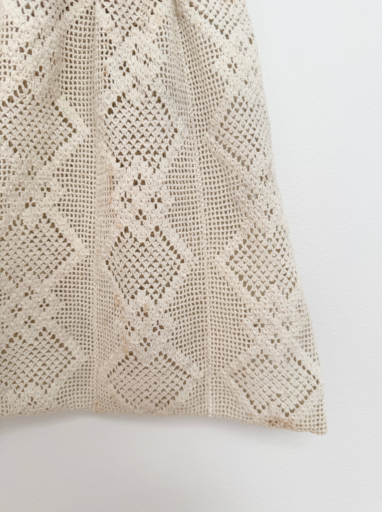 A close-up of a cream-colored crochet or lace fabric with a repeating diamond pattern, shown against a plain white background.