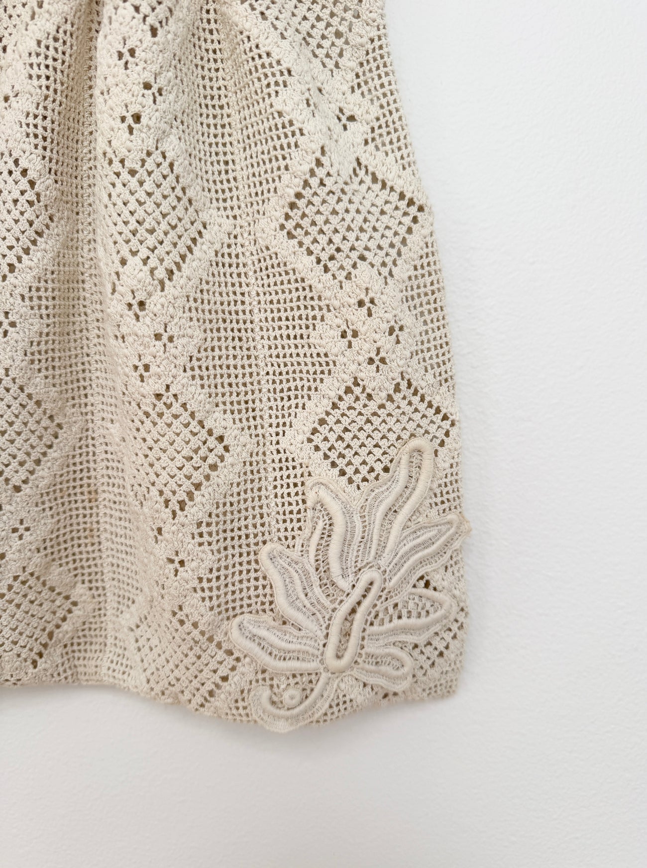 Close-up of a cream-colored lace garment, showing a detailed floral lace pattern and a matching embroidered applique on a plain white background.