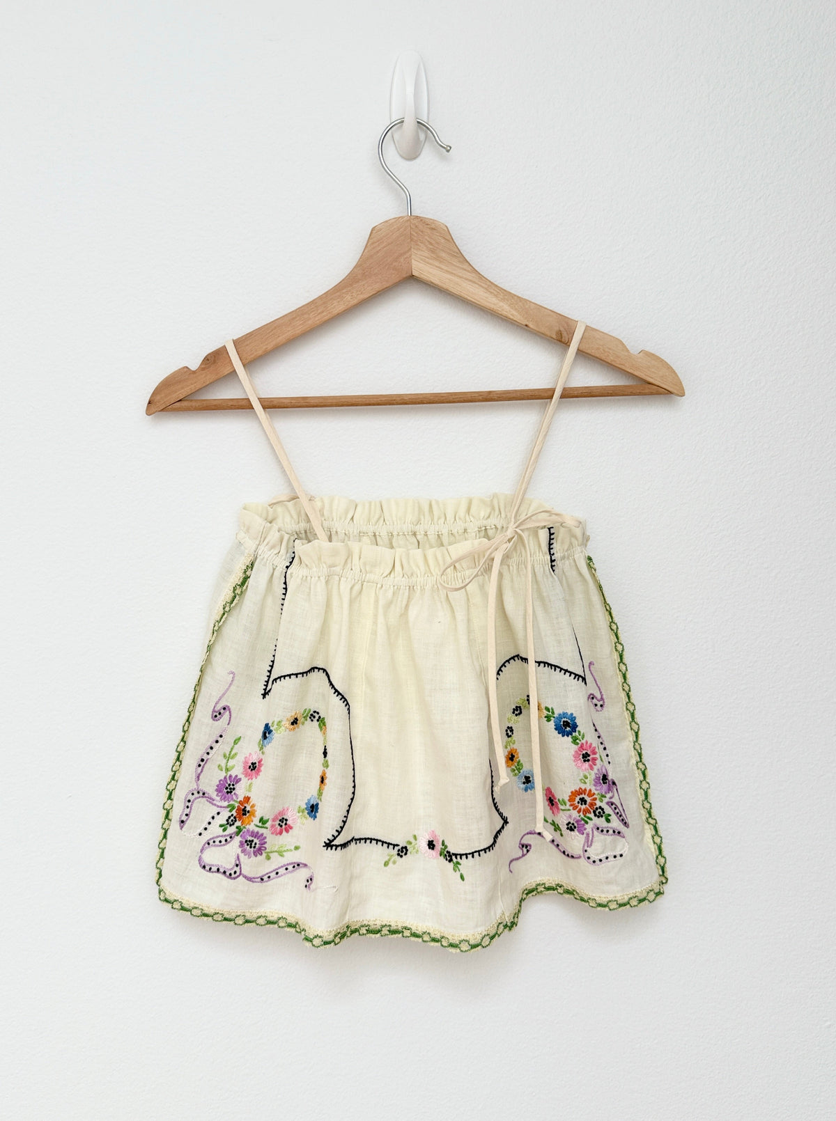 A cream-colored cotton camisole top with colorful floral embroidery and green trim, hanging on a wooden hanger against a plain white wall.