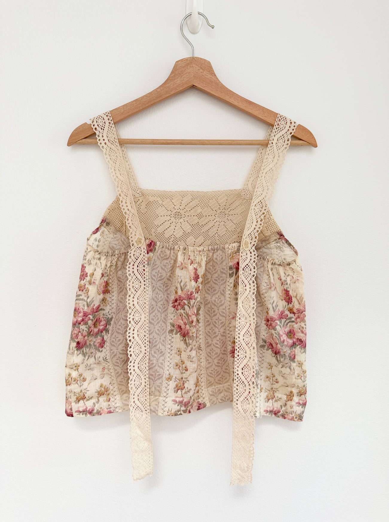 A sleeveless floral lace top in beige, featuring wide shoulder straps with lace trim and tie details, hanging on a wooden hanger against a plain white wall.
