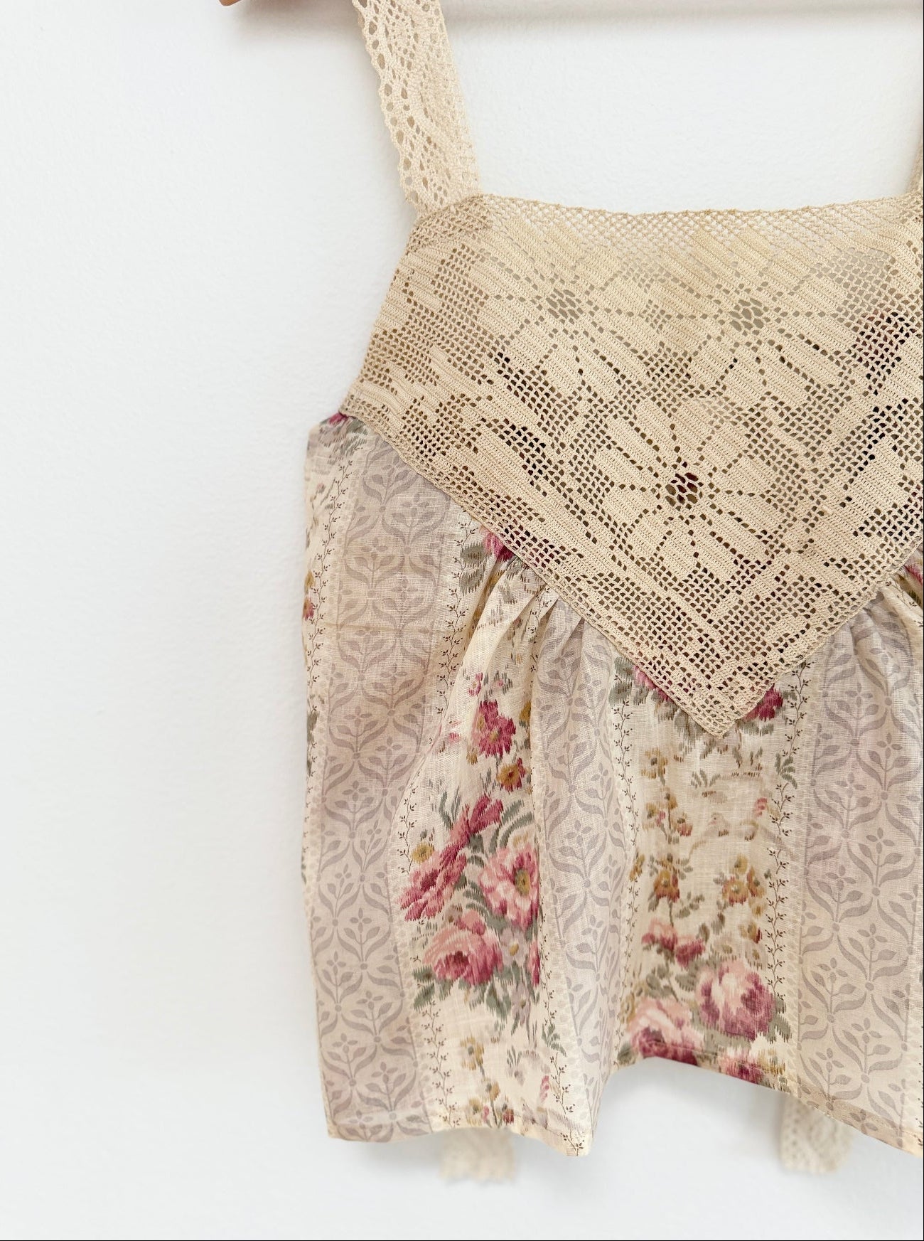A close-up of a cream-colored lace and floral fabric garment, showing a floral lace bodice overlay and patterned skirt panels, hanging against a plain white background.