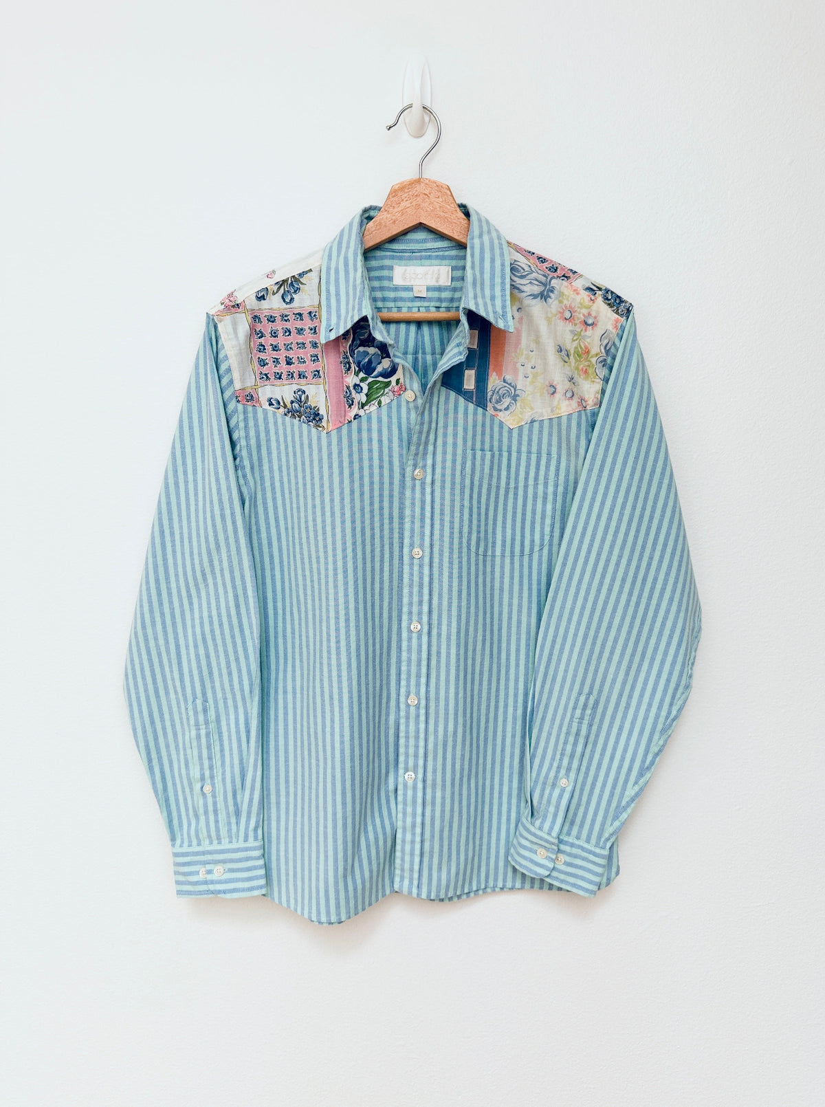 A light blue and white vertically striped long-sleeve button-up shirt with multicolored floral and patterned yoke panels on the shoulders, hanging on a wooden hanger against a plain off-white wall.