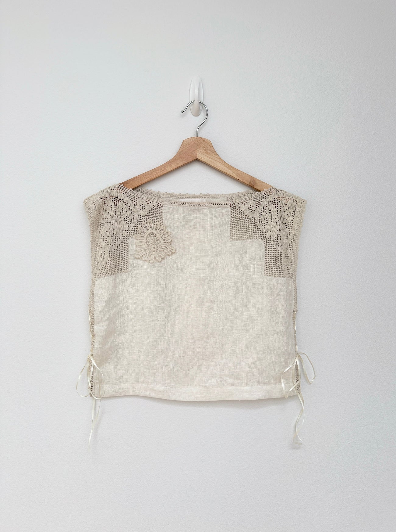 An off-the-shoulder cream linen top with beige crochet lace detailing on the chest and side ties, hanging on a wooden hanger against a light gray wall.