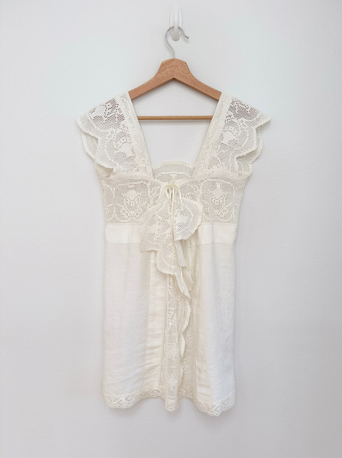 An off-white cotton crochet dress hanging on a wooden hanger against a plain white wall.