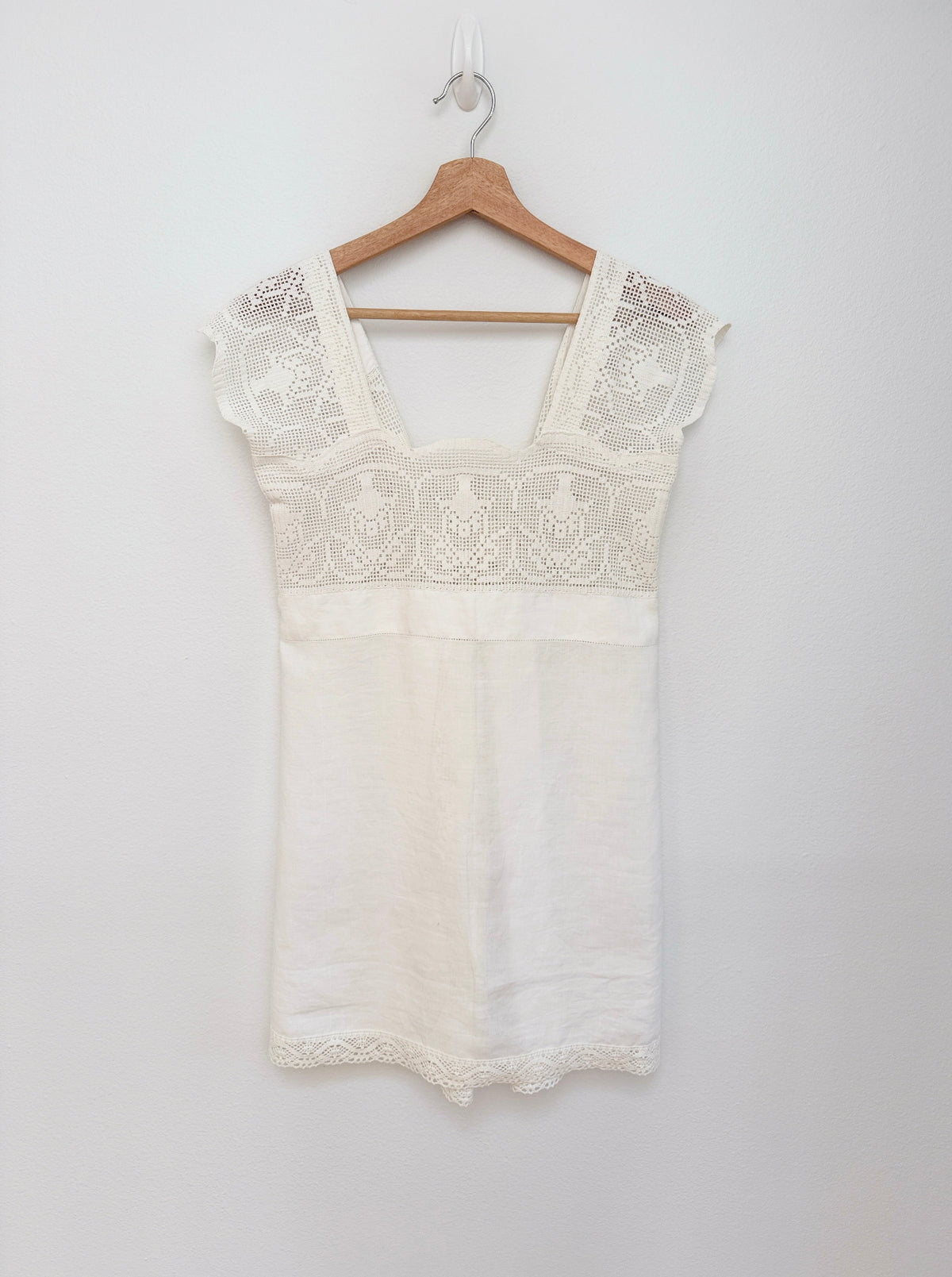 An off-white cotton dress with floral crochet trim on the bodice and cap sleeves, hanging on a wooden hanger against a plain white wall.