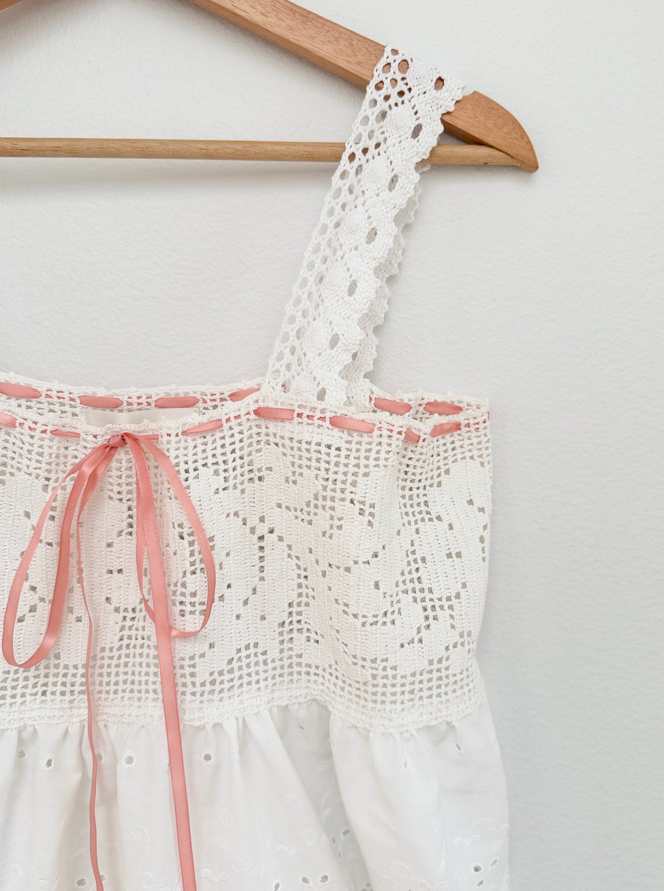 White eyelet lace top with pink ribbon trim and tie, hanging on a wooden hanger against a light wall.
