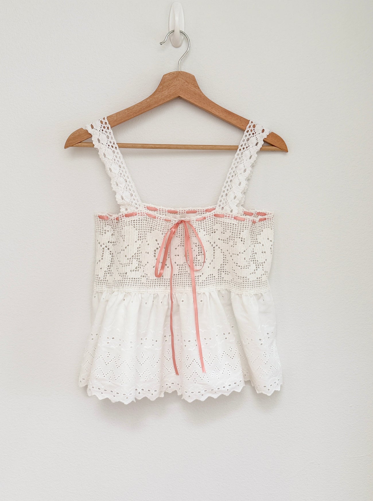 A white eyelet crochet sleeveless top with a pink ribbon tie at the center, hanging on a wooden hanger against a plain white wall.