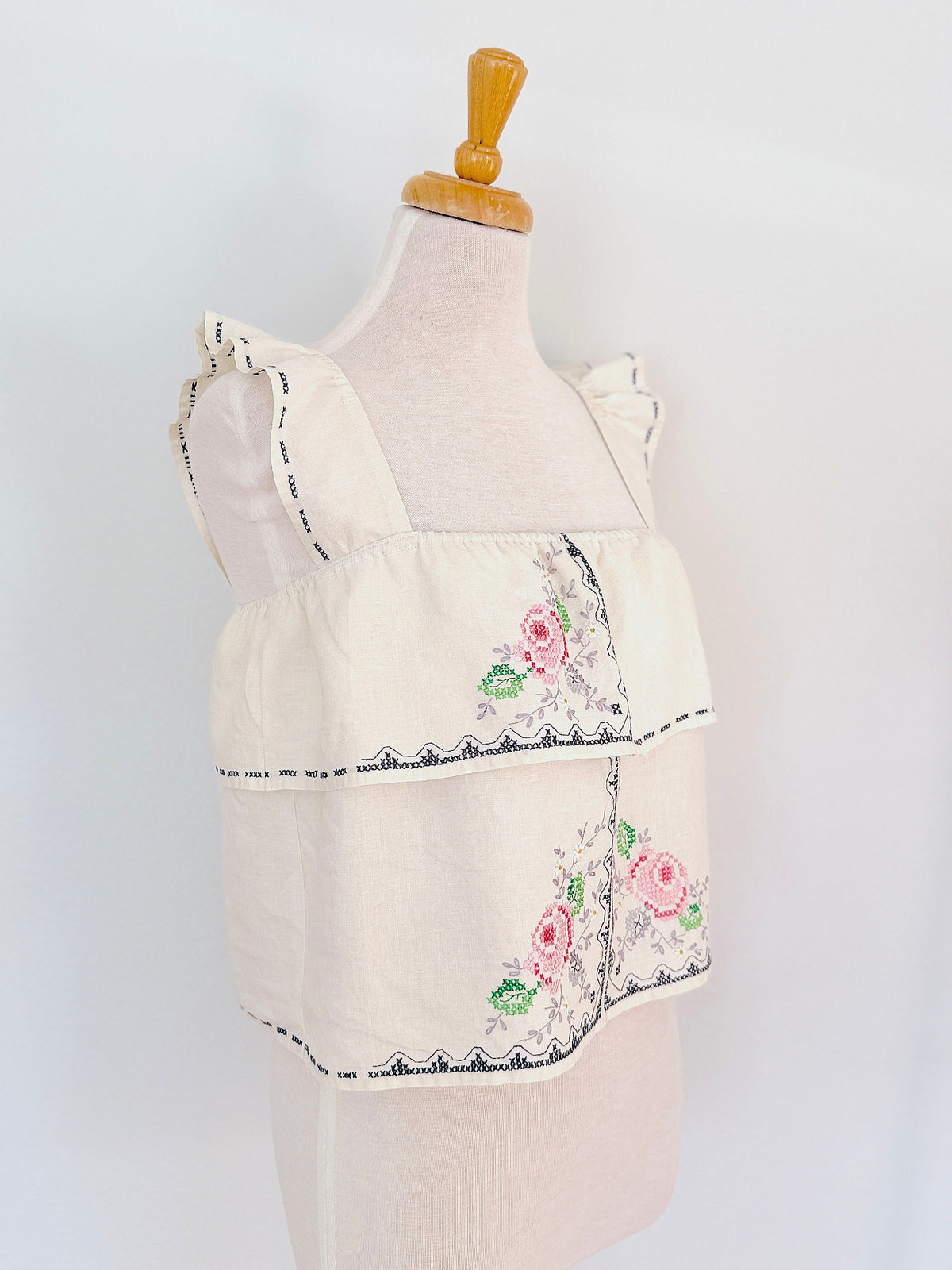 An off-white sleeveless blouse with ruffled shoulder straps, tiered ruffle layers, and pink floral embroidery with green leaves, displayed on a cream-colored dress form mannequin against a white background.