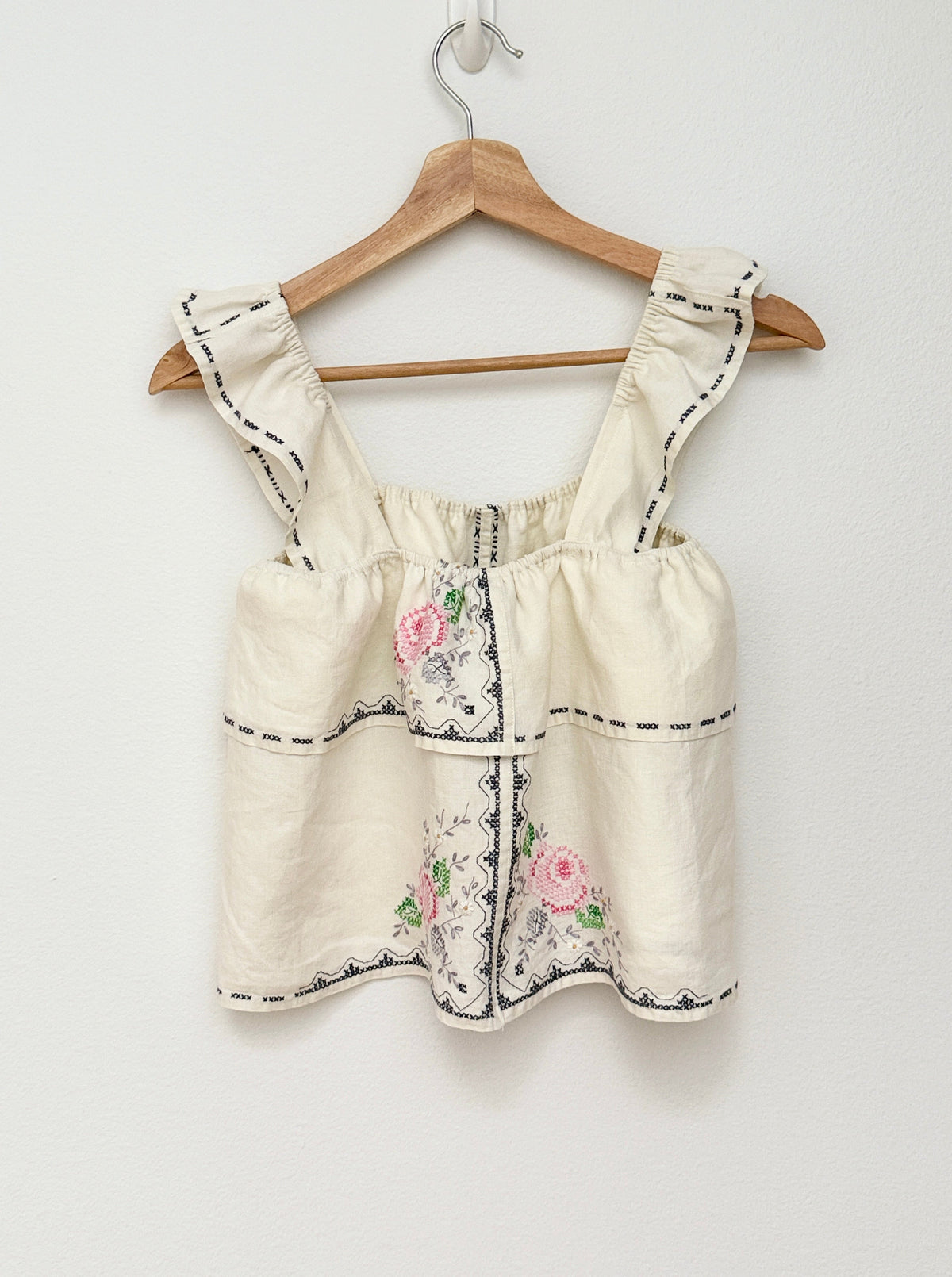 An off-white sleeveless top with floral embroidery and black stitched trim, hanging on a wooden hanger against a plain white wall.